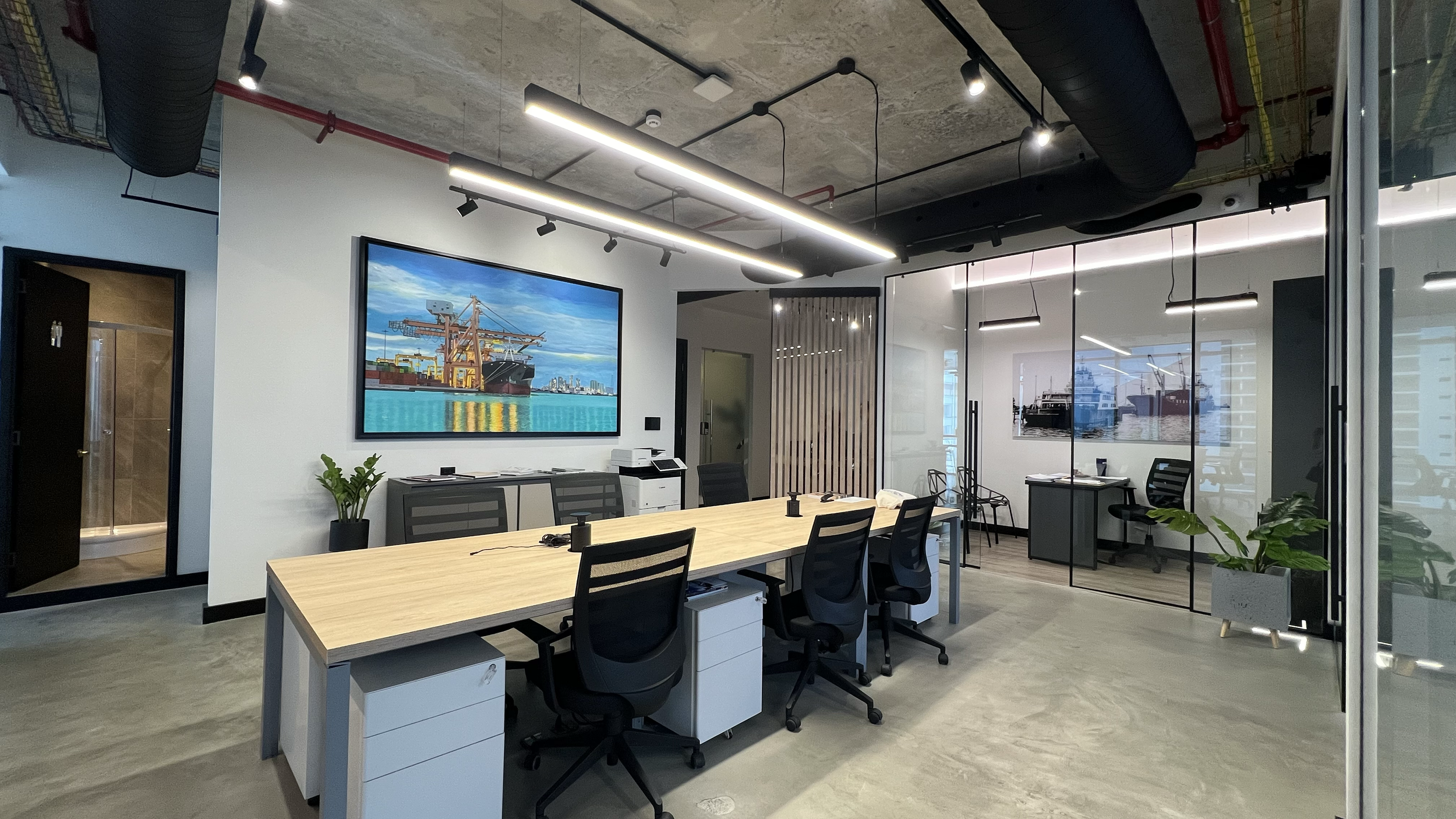 Modern office conference room with a long table, black chairs, large artwork of an oil rig and a ship, glass walls showcasing a maritime scene, ceiling with exposed concrete and industrial lighting, and potted plants.