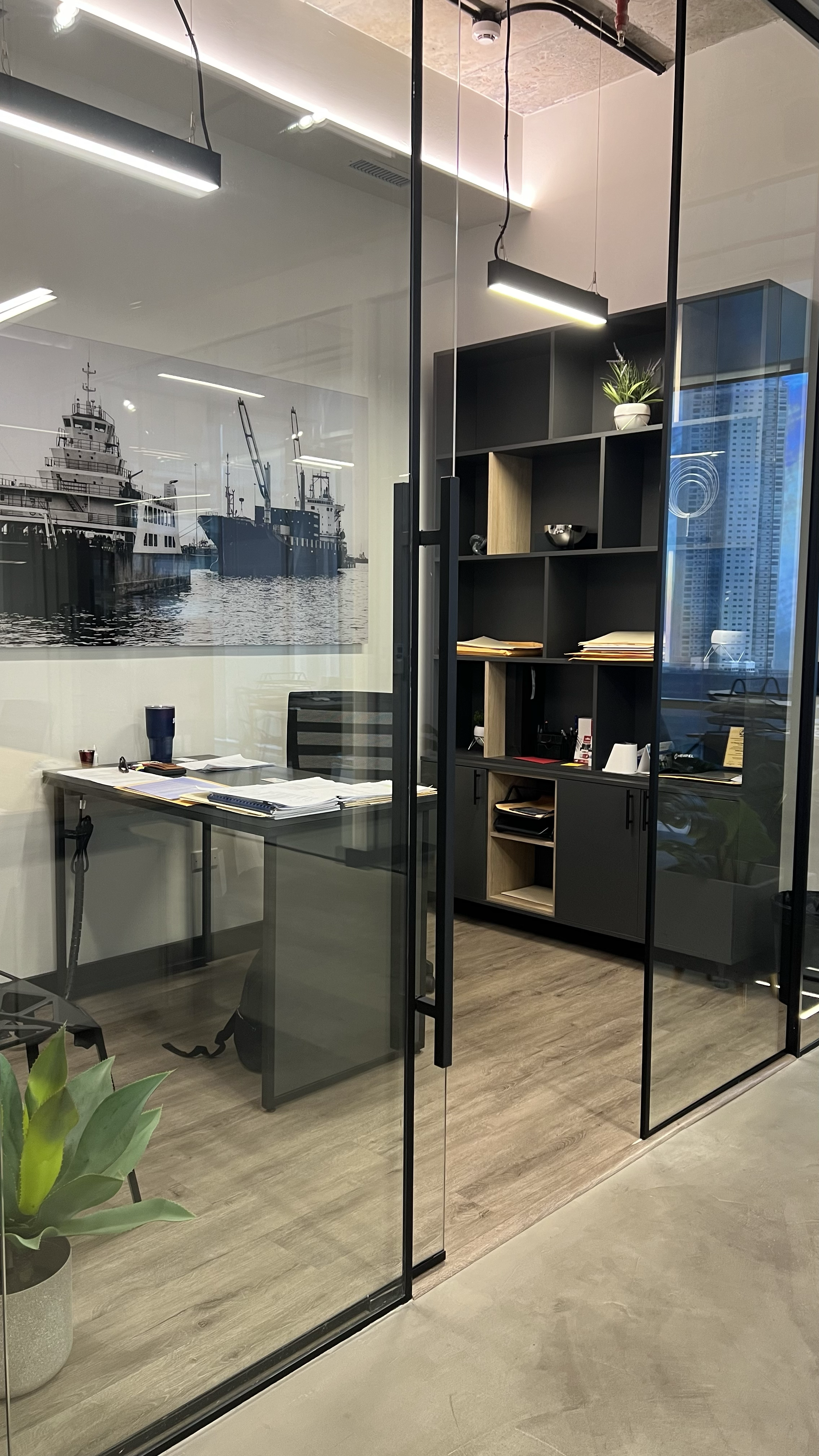 Inside an office with a glass wall, a desk with papers and a water bottle, a black swivel chair, a black storage cabinet with books and documents, a large maritime-themed black and white photo on the wall, and a potted plant in the foreground.