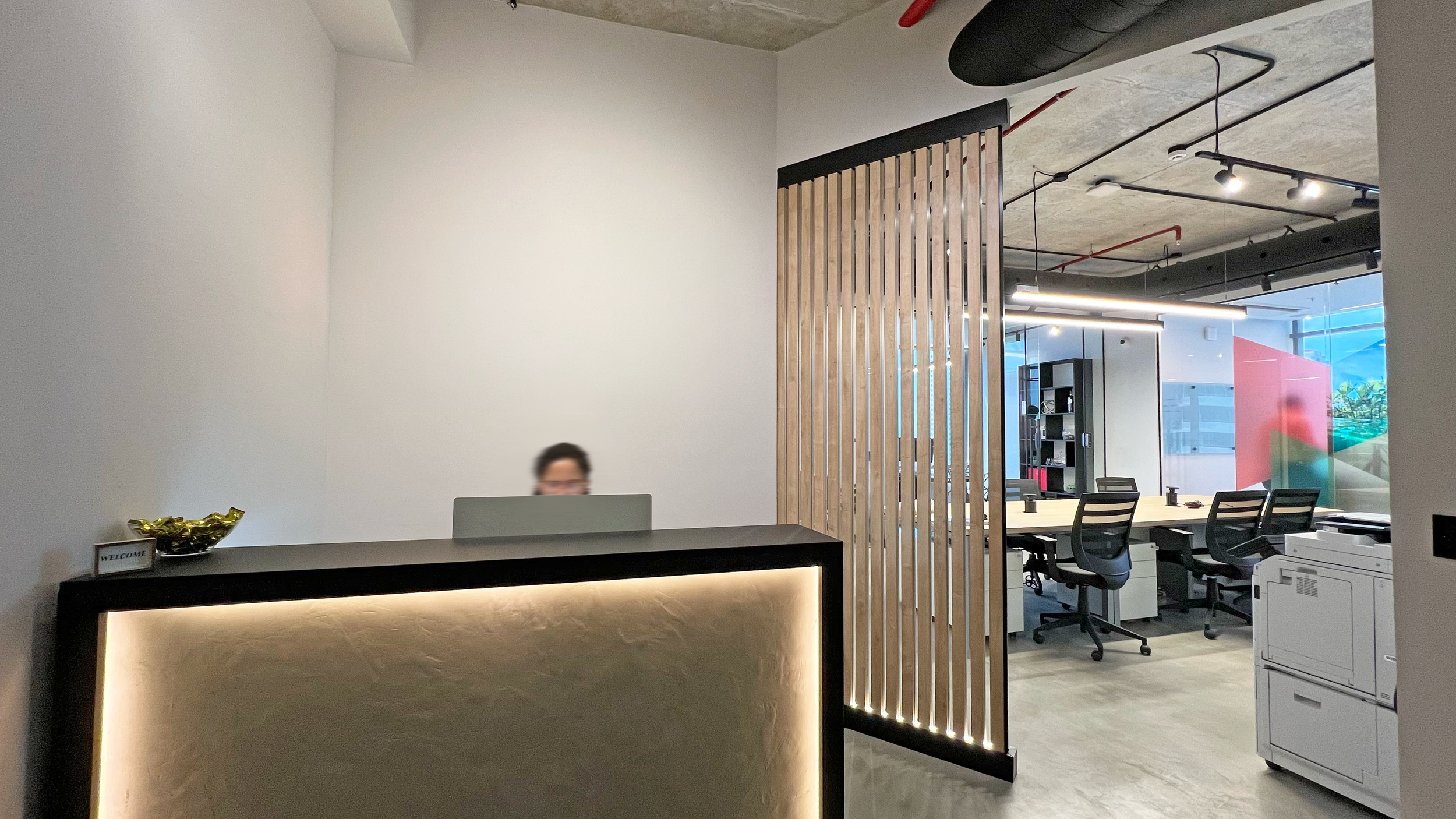 Modern office reception area with a person behind the reception desk, a decorative bowl, and a wooden slat partition. In the background are open workspaces with black office chairs, a large window, and ceiling lighting.