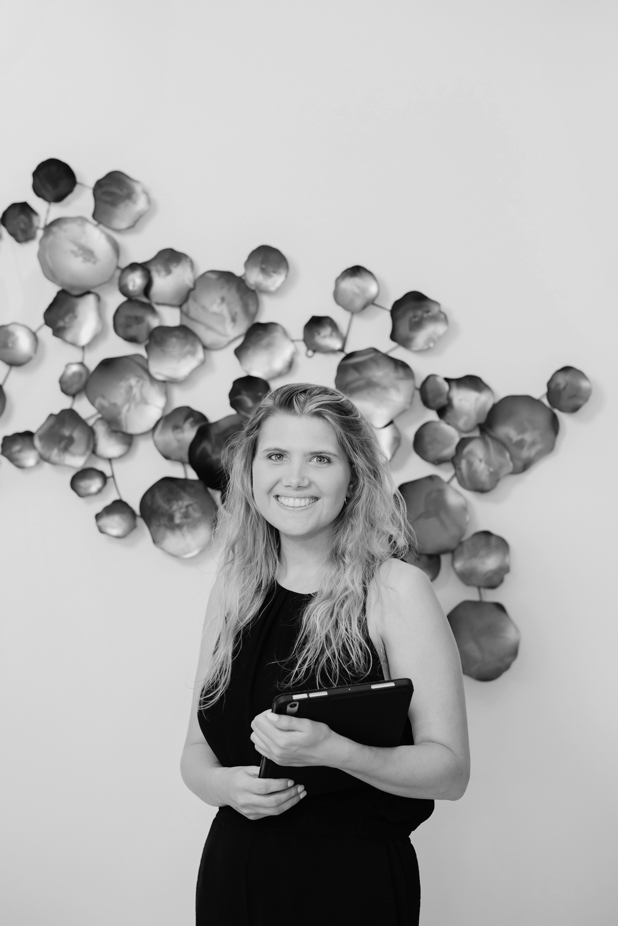 A woman with long wavy hair, wearing a sleeveless black dress, holding a tablet, and smiling in front of an abstract wall sculpture with rounded, metallic-looking shapes.