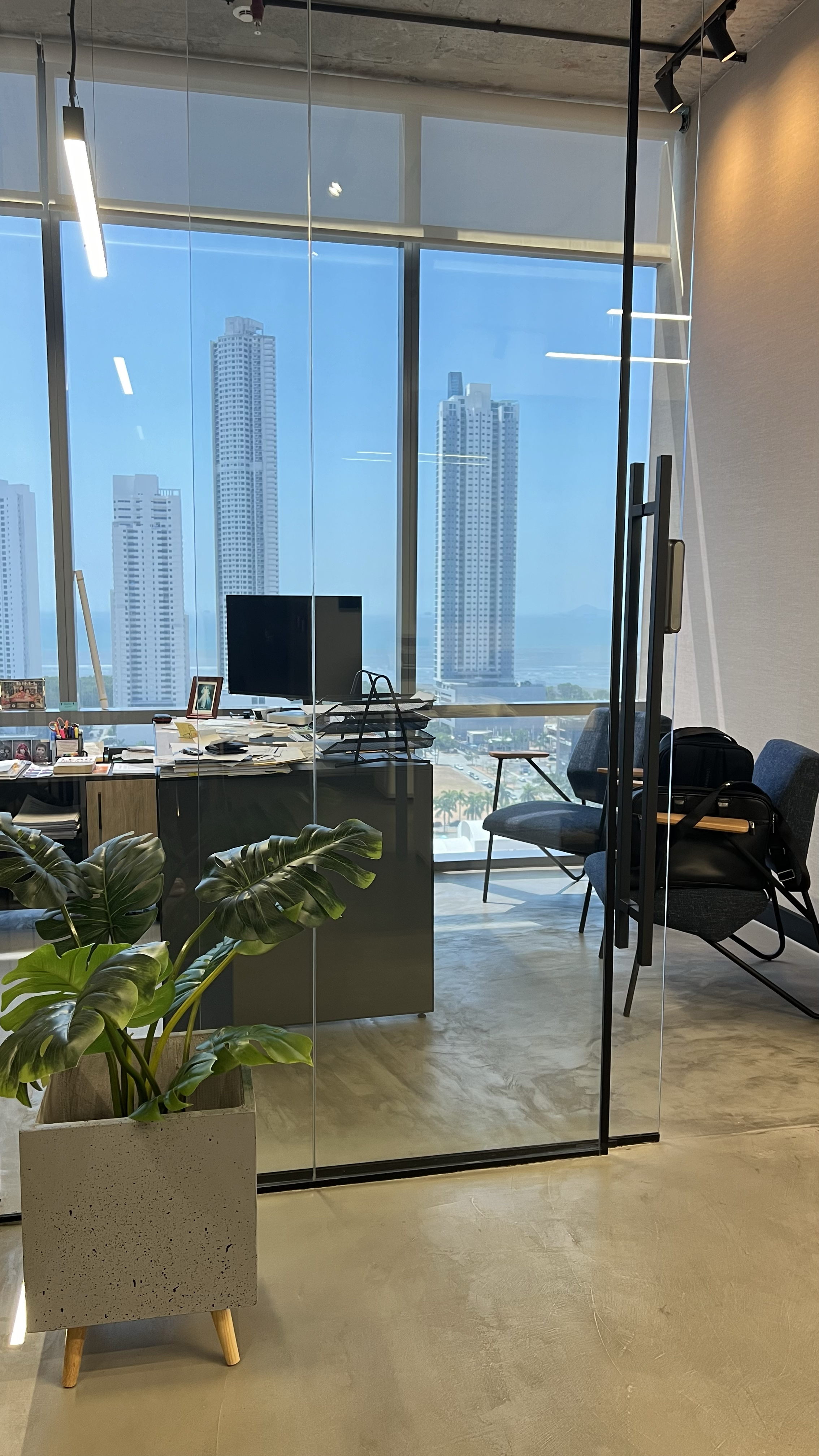 Office space with large glass windows showing skyscrapers and cityscape outside, desk with cluttered papers, computer monitor, and office chair, plant in the foreground, and additional chairs near the window.
