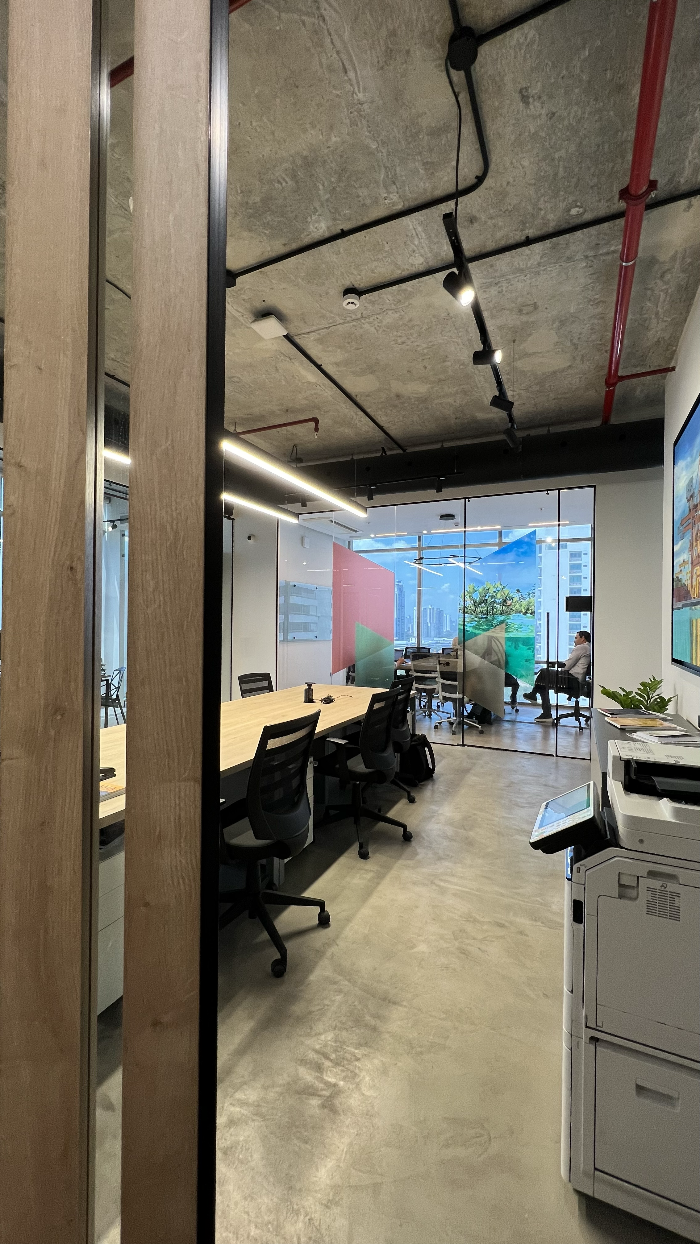 A modern office conference room with a long wooden table, black chairs, large windows with city views, and colorful geometric art on the windows. There is a person sitting by the window and office equipment like a printer in the foreground.
