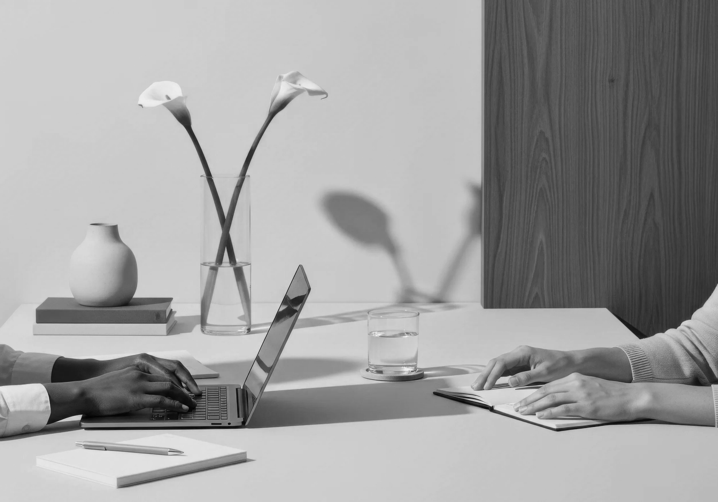 Black and white photograph of a meeting or workspace, showing two pairs of hands on a table with a laptop, a notebook, and a pen. In the background, there are decorative vases, a glass of water, and a wall with a wooden panel.