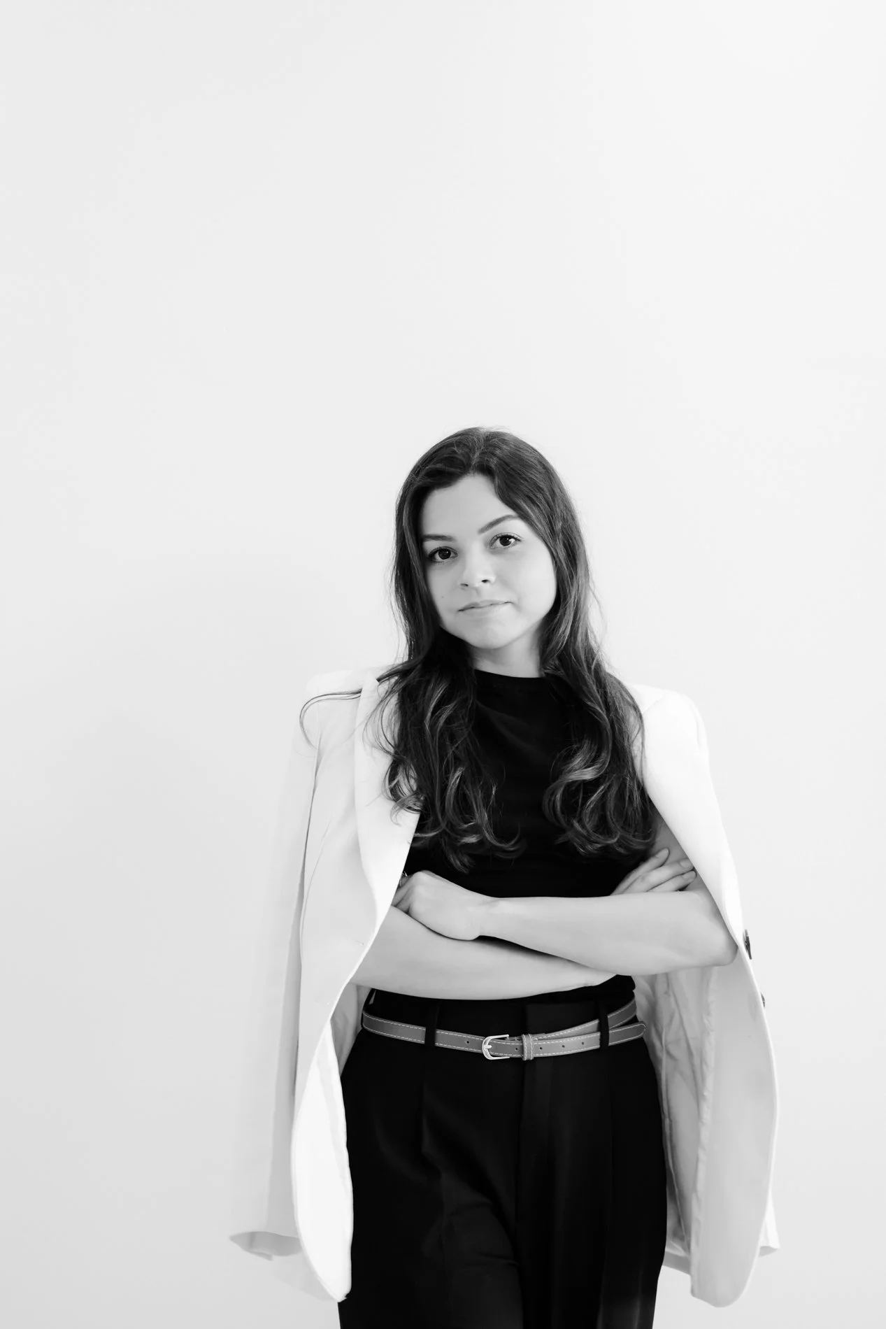 Black and white portrait of a woman with long wavy hair, crossed arms, wearing a white blazer over a black top, standing against a plain light background.