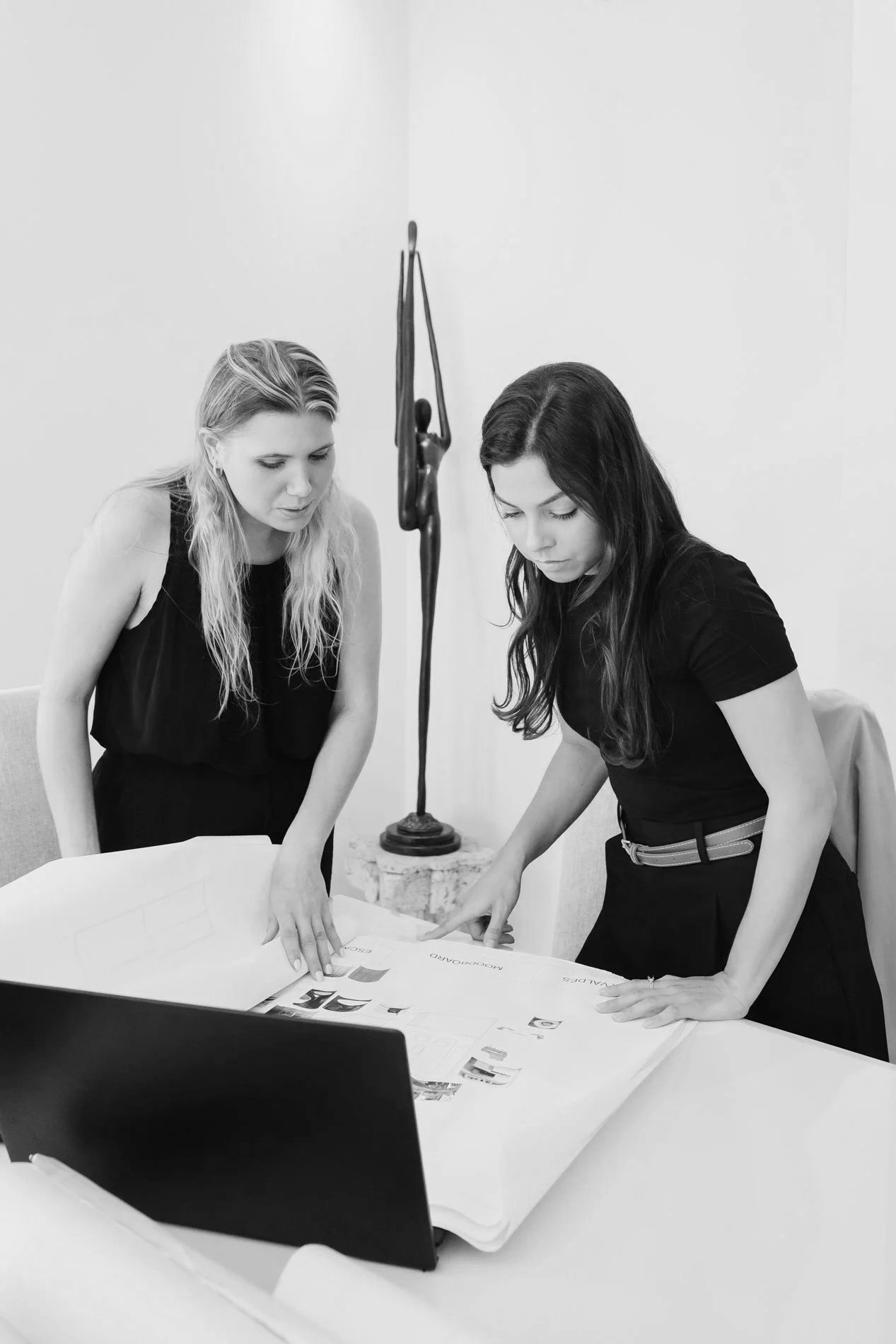 Two women leaning over a table, looking at a large open presentation board with images and text, in a meeting room with white walls and a sculpture in the background.