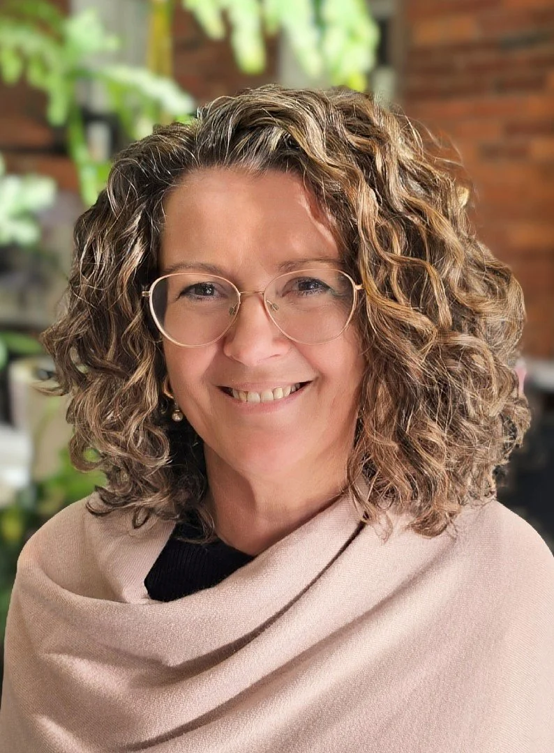 A woman with curly brown hair wearing glasses and a beige shawl, smiling with a brick wall and green plants in the background.