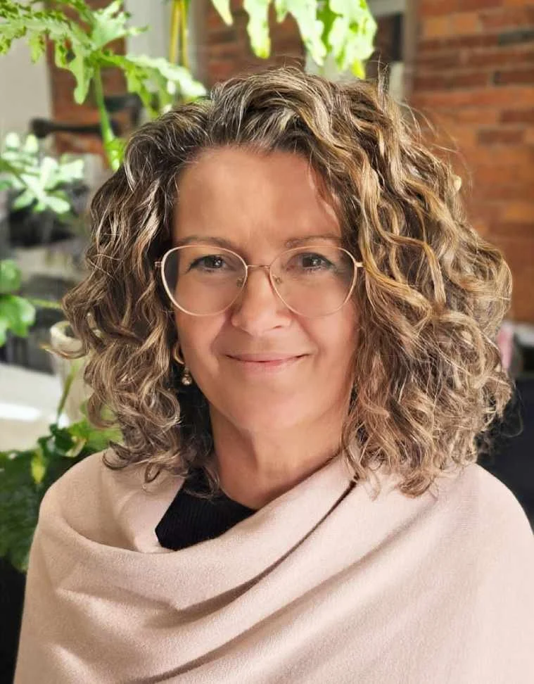 A woman with curly hair, wearing glasses, and a beige shawl, smiling indoors with green plants and a brick wall in the background.