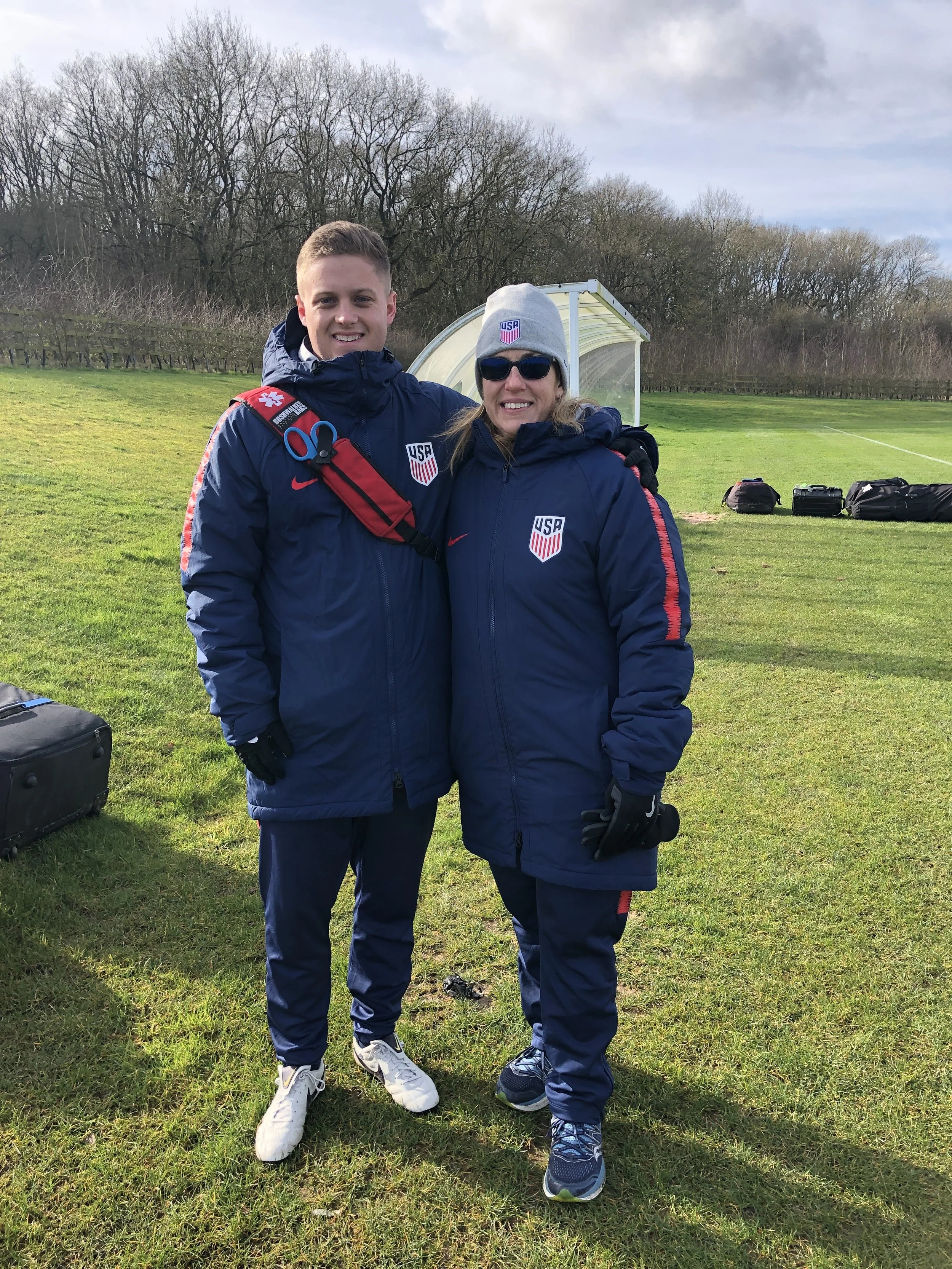 Two people in US national team jackets standing outdoors on a grassy field, smiling, with equipment and a small covered bench in the background, trees, and a cloudy sky after a performance recovery acl session