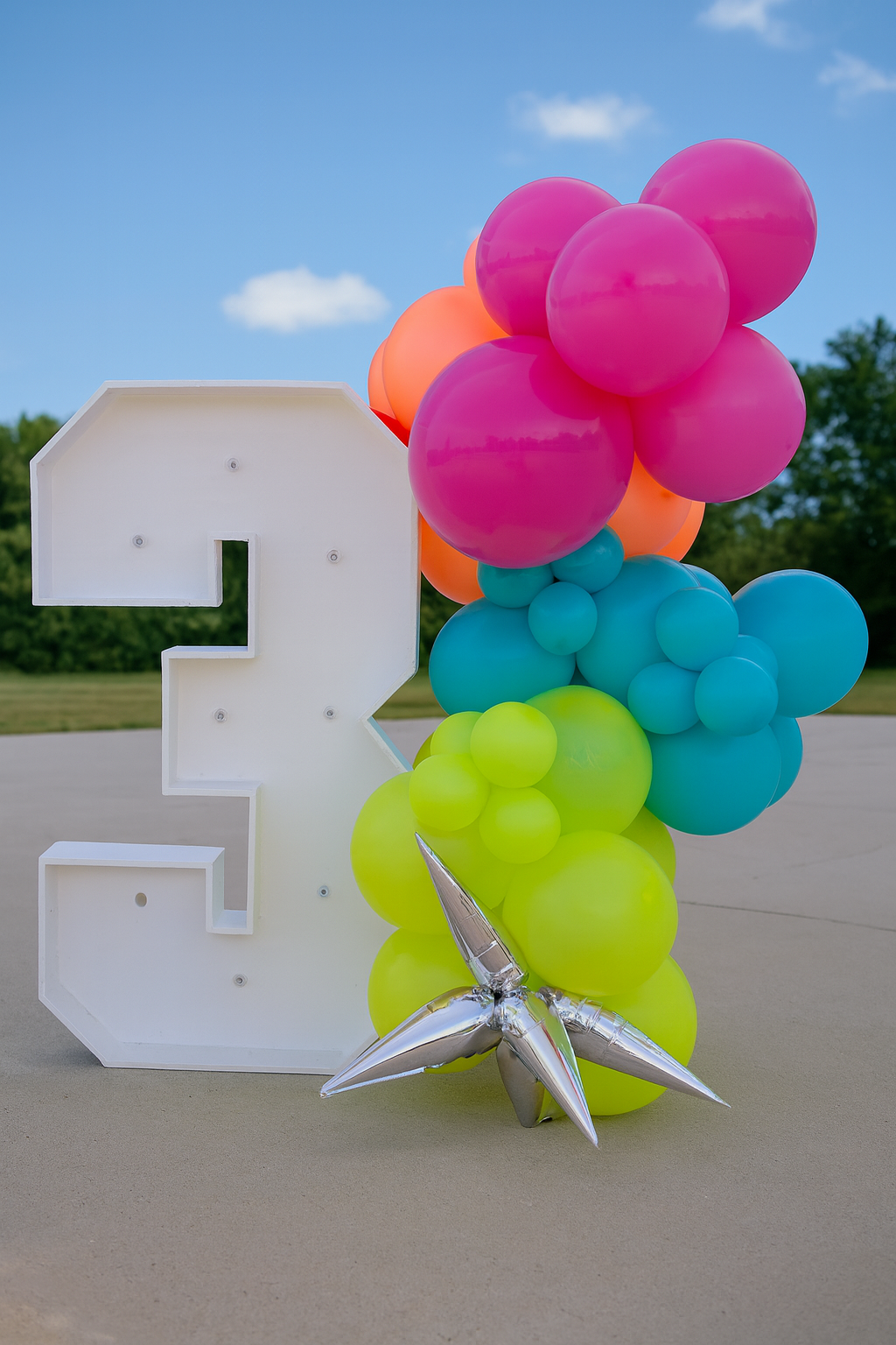 Large white number 2 decoration surrounded by colorful balloons including pink, orange, blue, and yellow, with a silver star-shaped balloon at the bottom, outdoors on a sunny day with a clear blue sky and trees in the background.