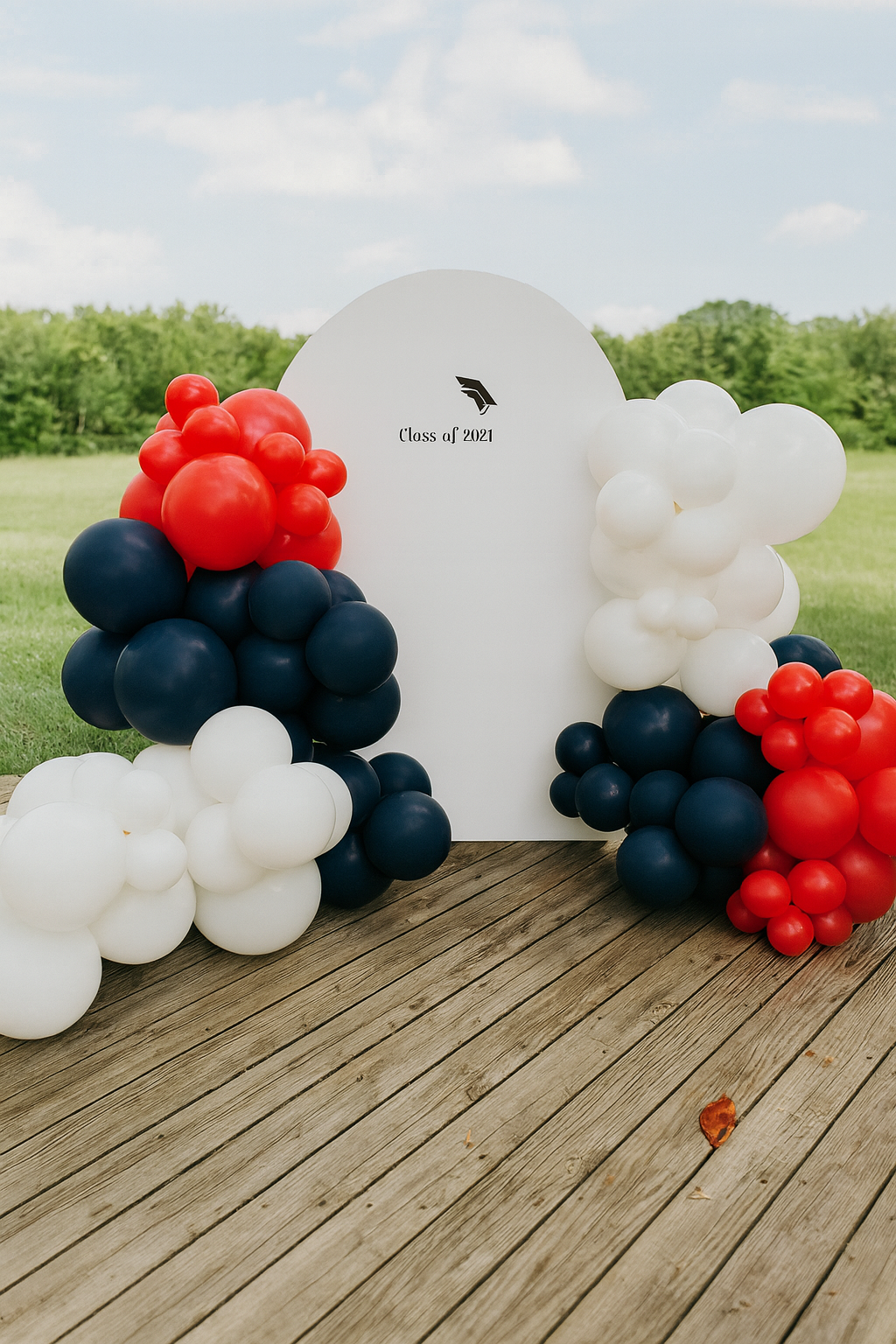 Graduation celebration backdrop with balloons in red, white, and navy blue on a wooden deck outdoors, with a grassy field and trees in the background, and a sign that reads "Class of 2021" with a graduation cap logo.