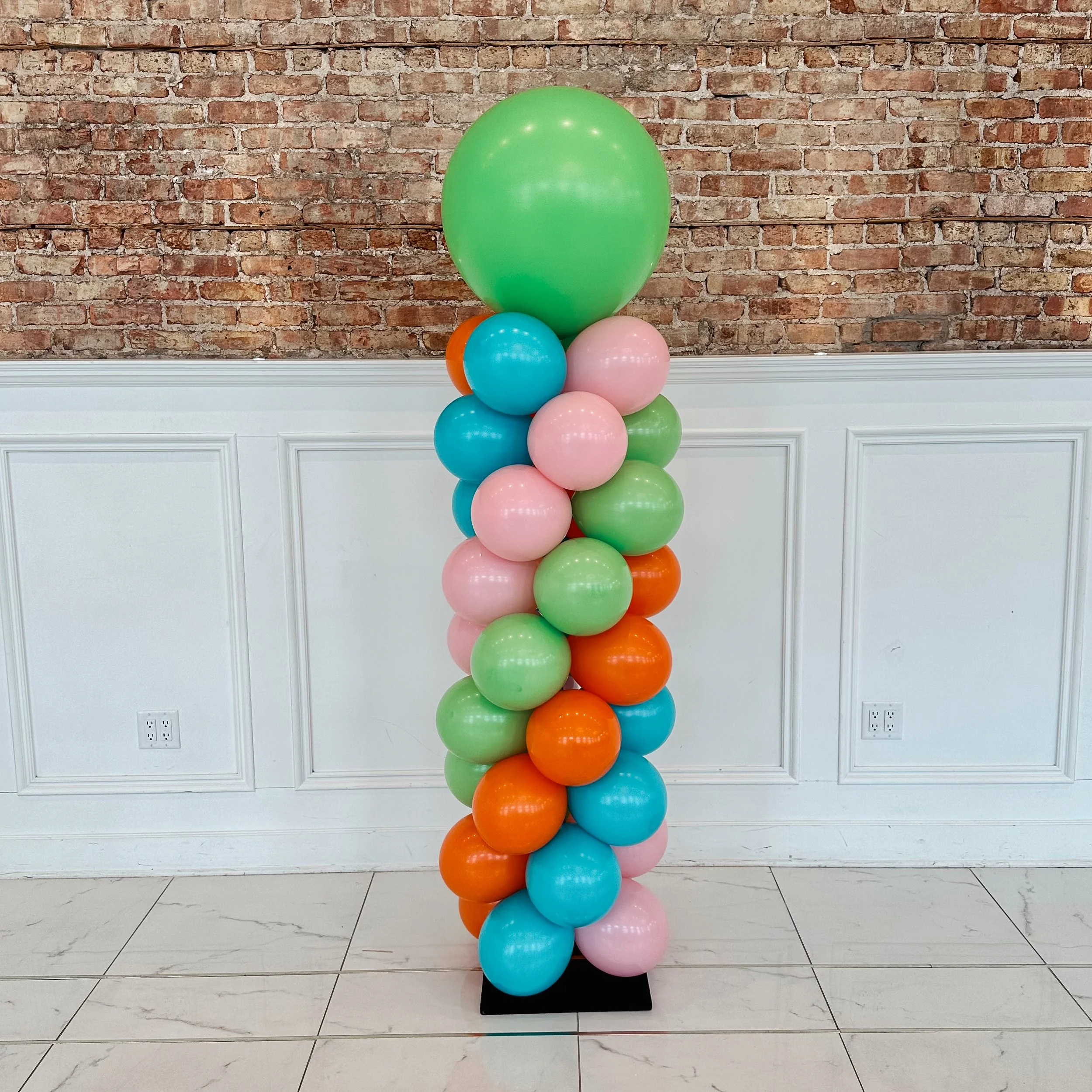 Colorful balloon arrangement with pink, blue, green, and orange balloons stacked vertically, standing on a black base against an exposed brick wall.