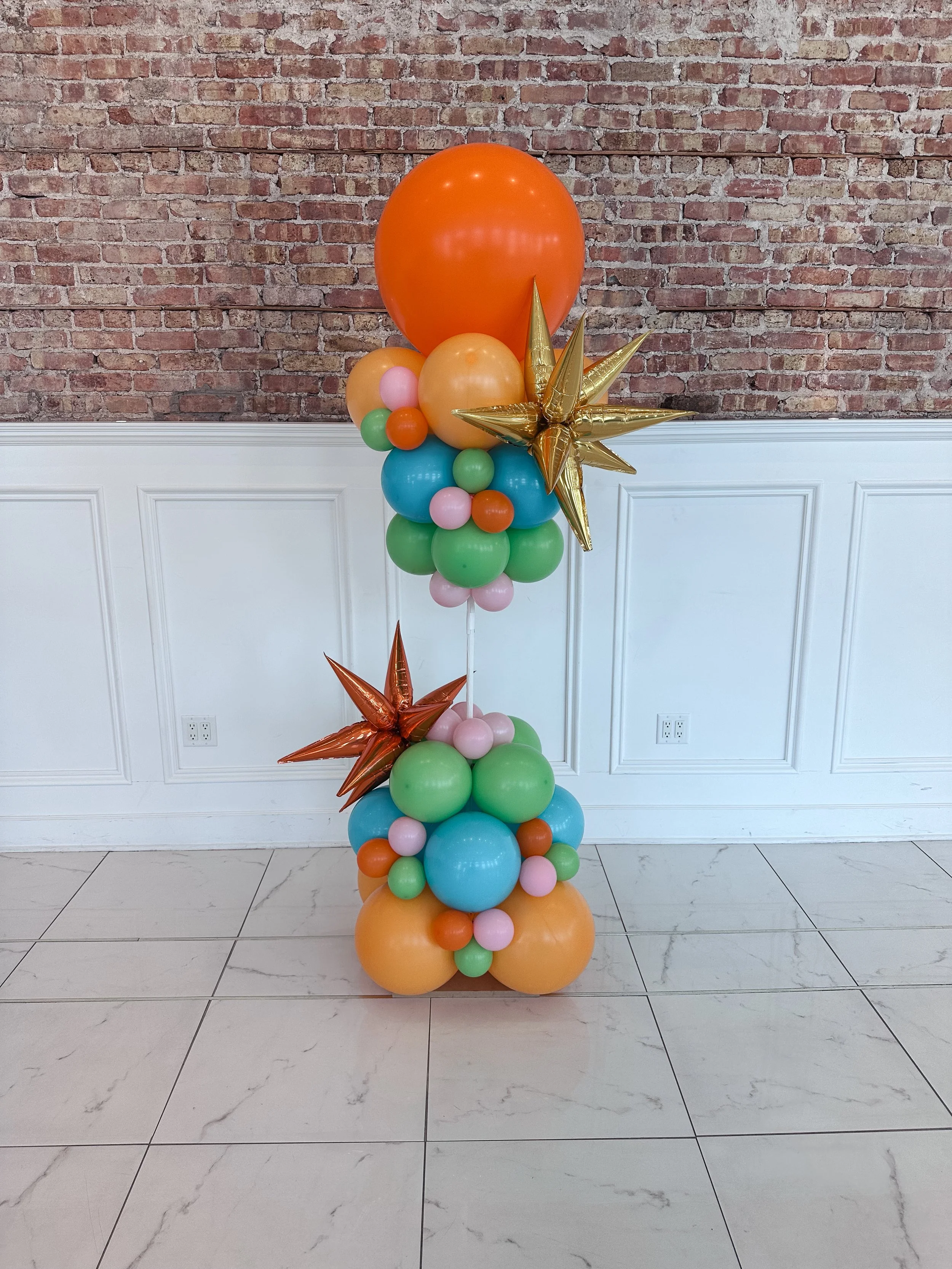 Colorful balloon arrangement with orange, yellow, pink, blue, green, and gold balloons, including star-shaped balloons, set against a brick wall background.