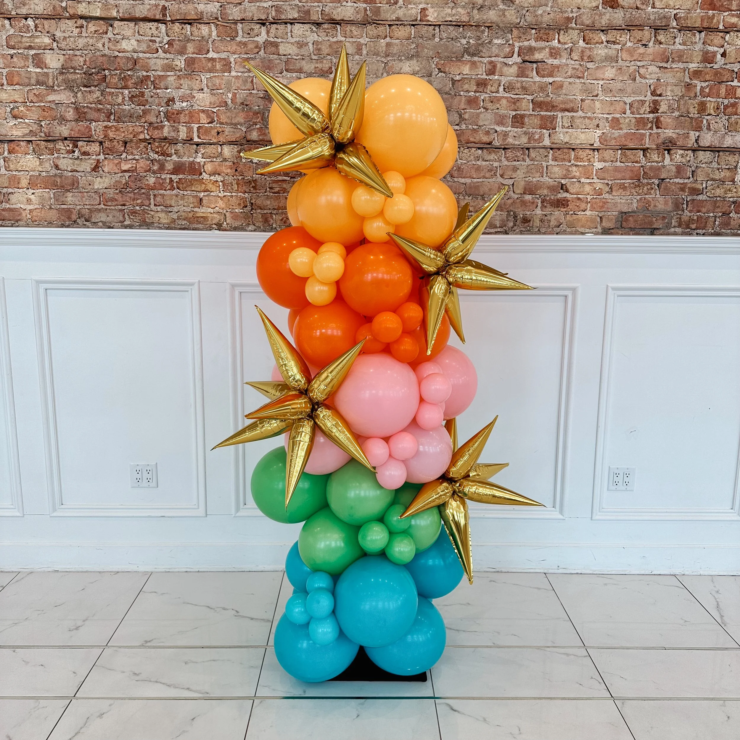 Colorful balloon arrangement with orange, pink, green, and blue balloons, accented with gold star-shaped balloons, set against a brick wall background.