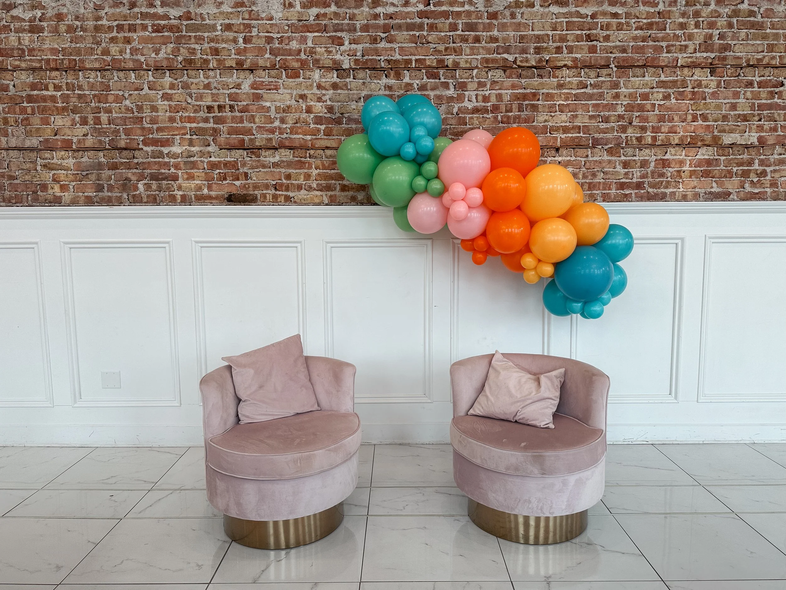 A wall decorated with pastel-colored balloons in pink, blue, green, orange, and yellow arranged in a cluster, with two pink velvet chairs and a brick wall background.