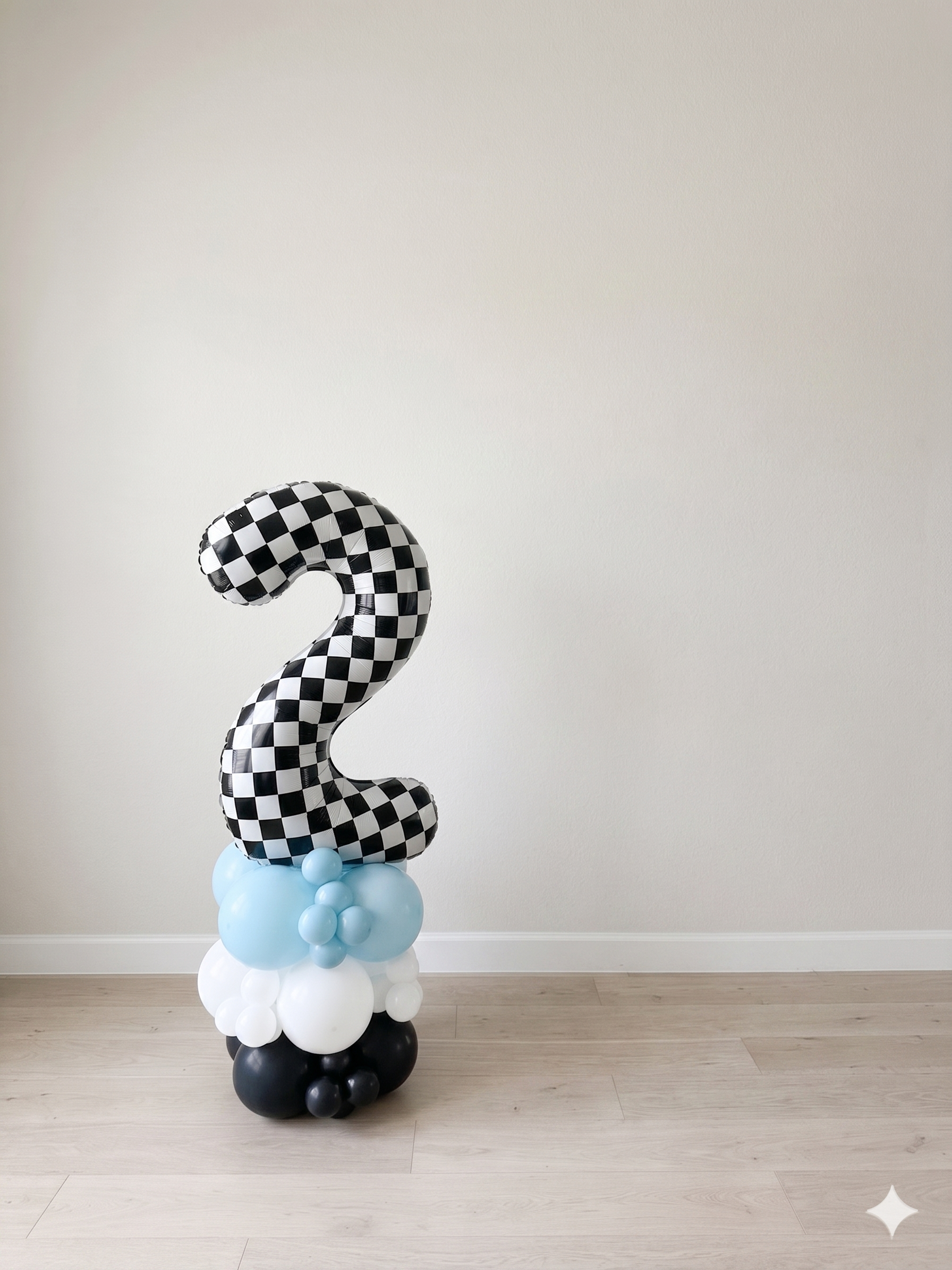 Balloon arrangement featuring a large black and white checkered number 2 on top of a cluster of white, light blue, and black balloons, some forming a cloud at the base, against a plain white wall.