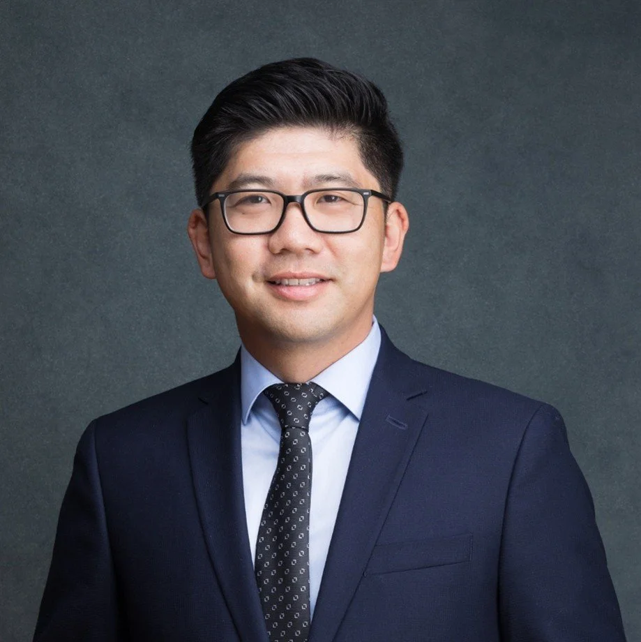 A professional man wearing glasses, a navy blue suit, light blue shirt, and a patterned black tie, smiling against a dark gray background.