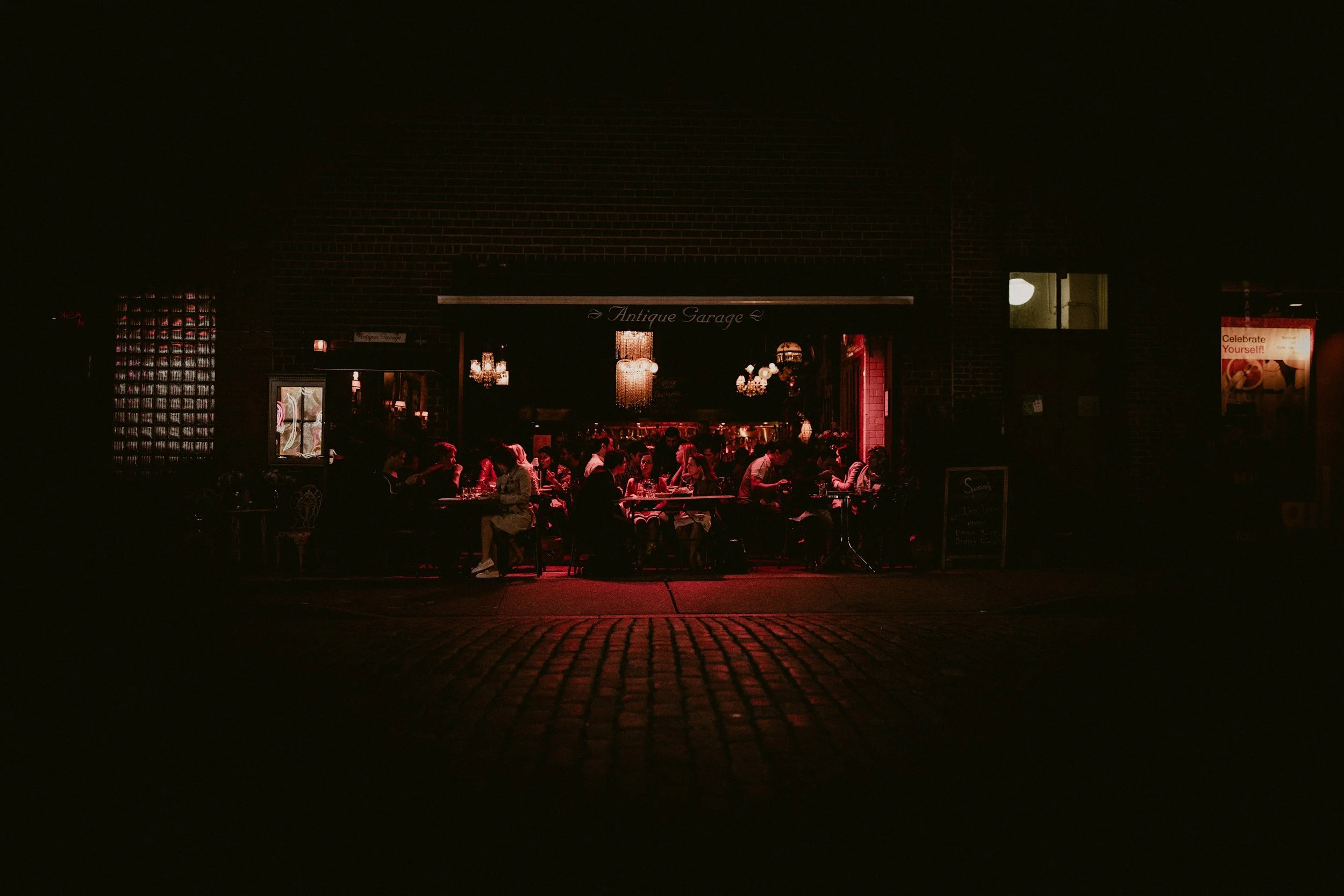 Night scene of a restaurant called 'Antique Garage' with people dining inside and outdoor patio, lit by warm hanging chandeliers and string lights, brick building facade, and a street sidewalk in front.