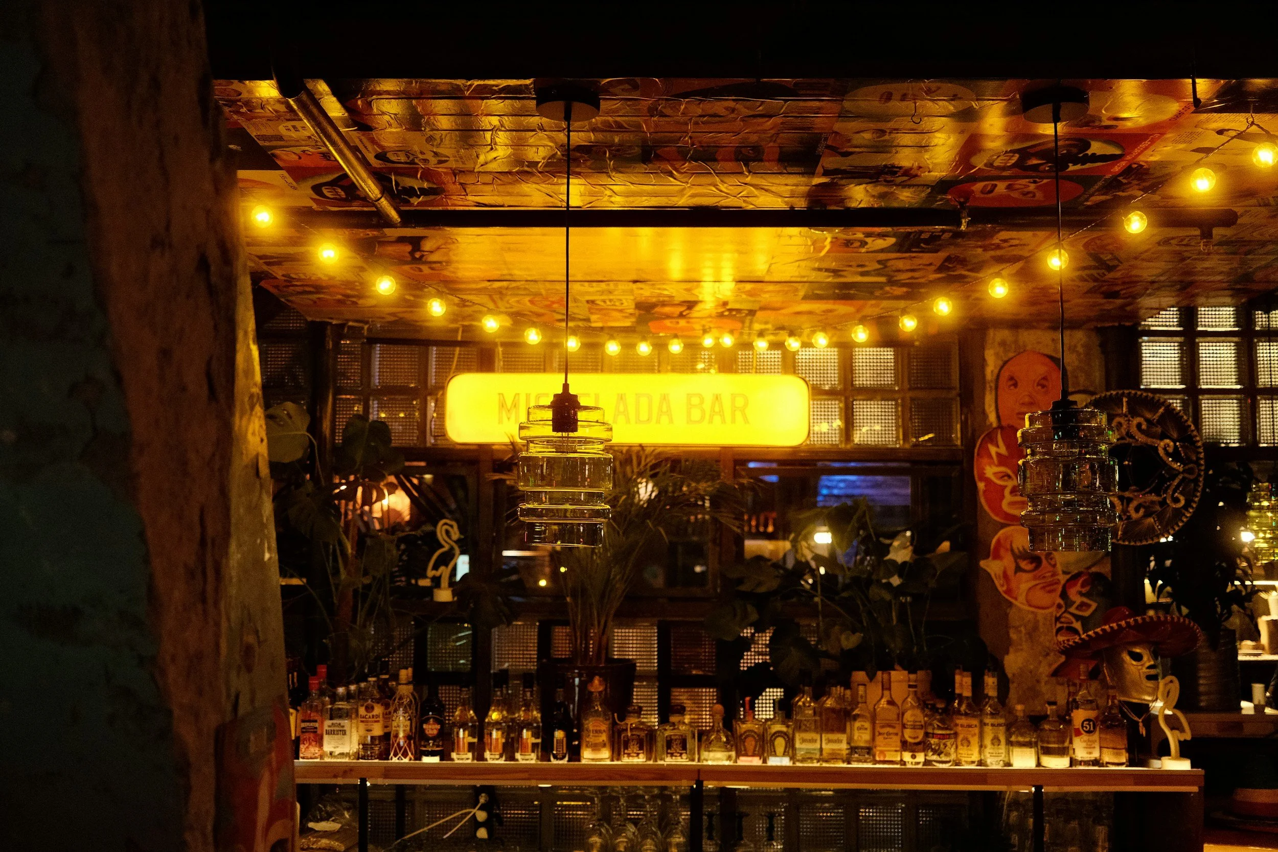 A bar with a bright yellow illuminated sign that reads 'MEXICANA BAR' in the background, with bottles of alcohol lined up on a shelf underneath. The ceiling is decorated with colorful posters or artwork, and warm yellow string lights are hanging, creating a cozy ambiance.