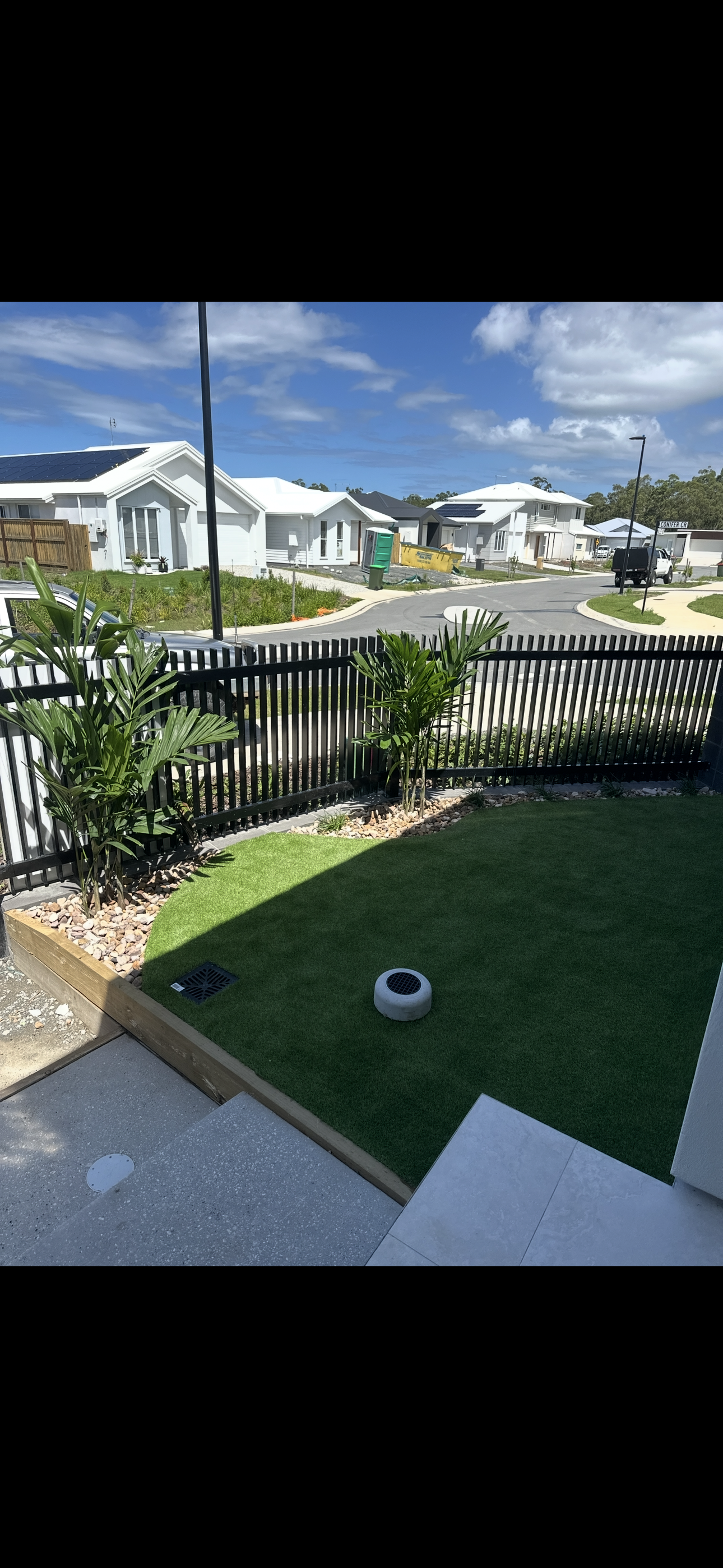 View from a backyard porch showing a well-maintained lawn, potted plants, a black fence, and a neighboring suburban street with modern houses, a clear blue sky, and some clouds.