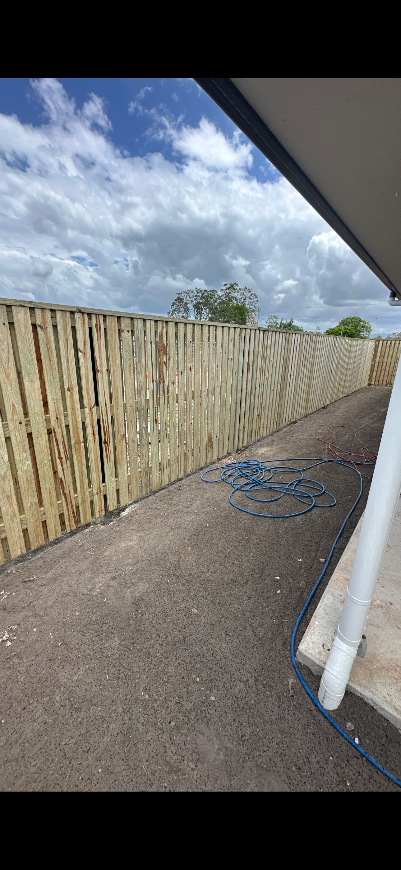 Backyard construction area with a newly installed wooden fence, a dirt ground, and some scattered garden hoses. Part of the house's roof and gutter are visible, with a partly cloudy sky overhead.