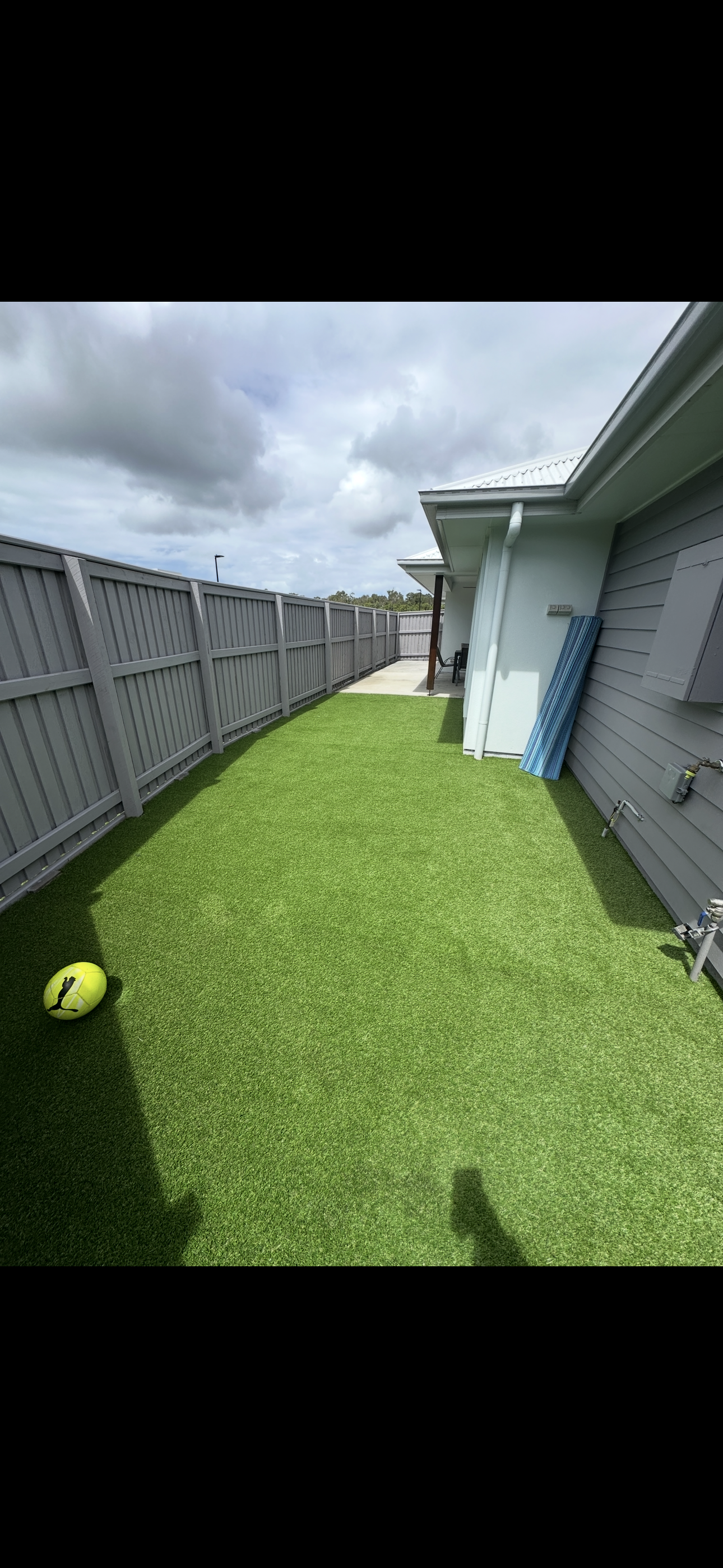 Backyard with artificial green grass, gray fencing, a house wall with an electrical box, a blue striped pool float leaning against the house, and a yellow tennis ball on the ground under cloudy sky.