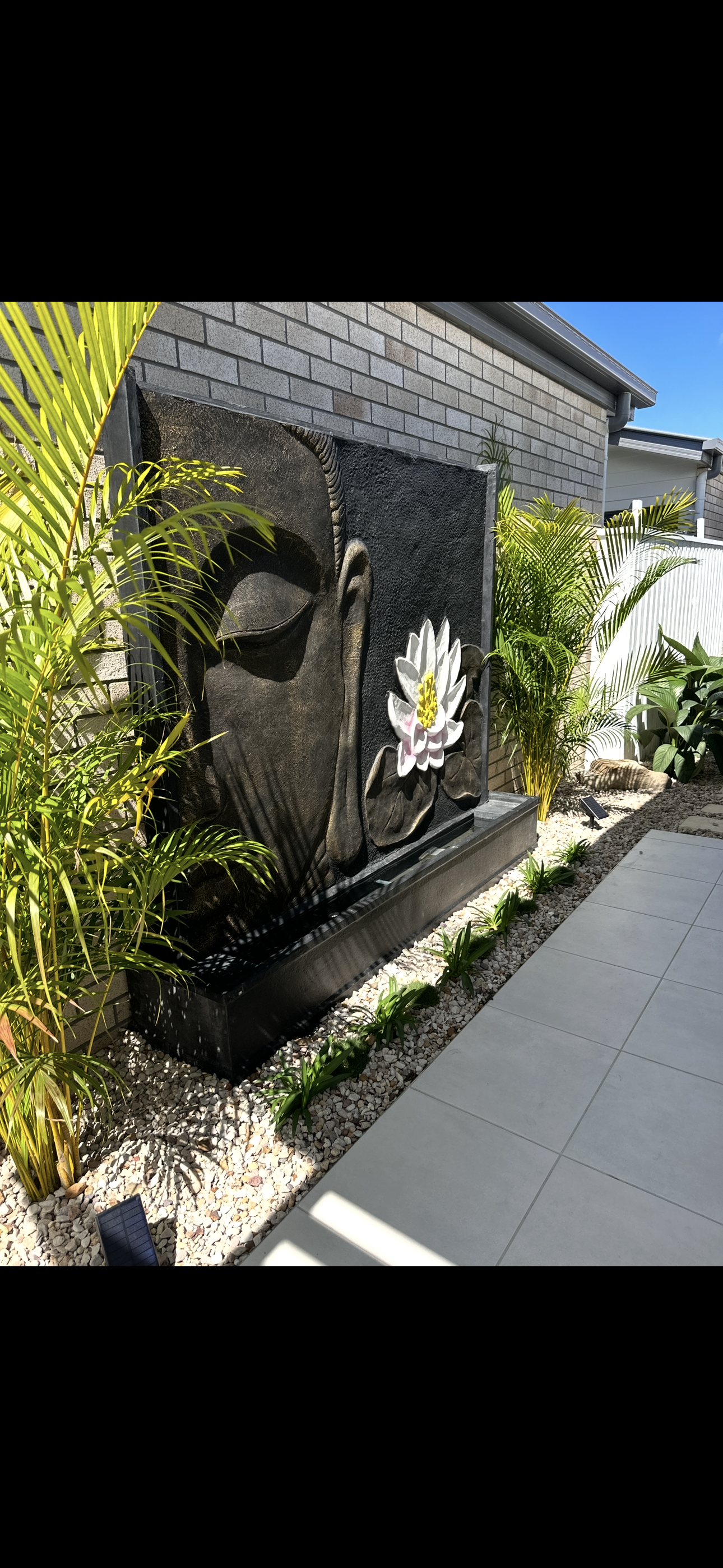 A garden water fountain sculpture of a Buddha head with closed eyes, surrounded by a white lotus flower, set against a brick wall with lush green plants and small white pebbles.