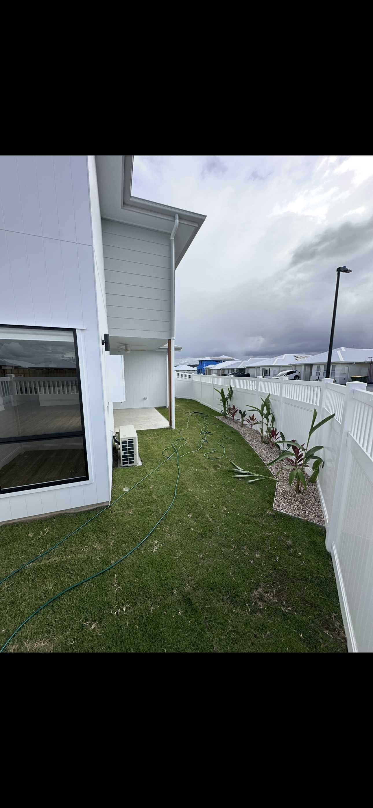 A small backyard with green grass, a white fence, and some young plants. There is an air conditioning unit and garden hoses on the ground. The house exterior is white. Overcast sky in the background.