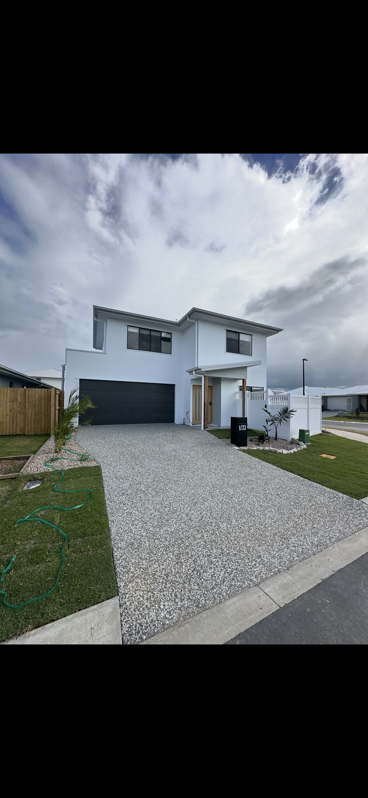 Modern two-story house with white exterior, black garage door, wooden details, and a pebble-textured driveway. Small landscaped yard with palm plants and a white fence.