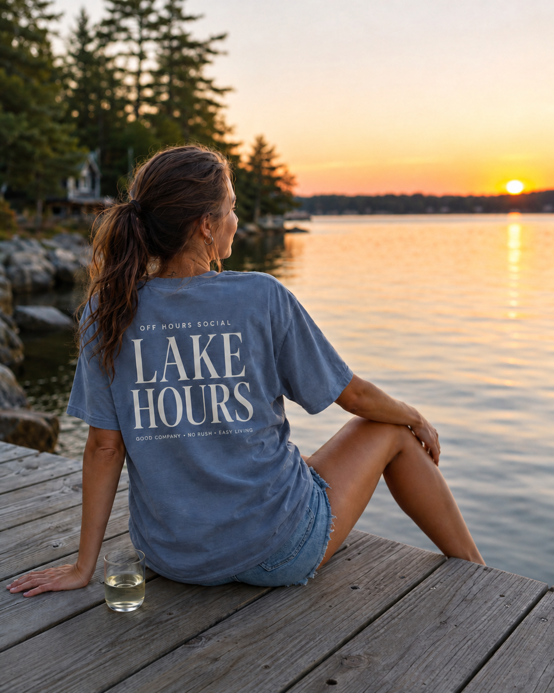 Woman sitting on a wooden dock by a lake during sunset, wearing a blue T-shirt with 'Lake Hours' written on the back, and a glass of white wine beside her.