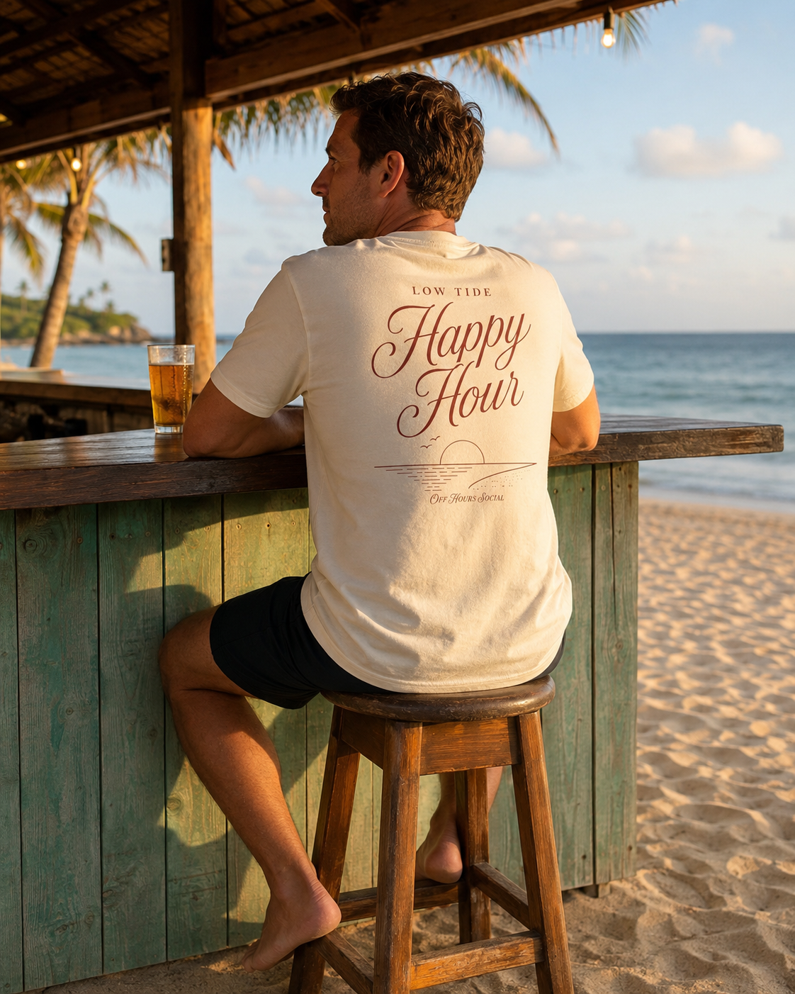 Man sitting on a wooden stool at a beach bar, facing the ocean, with a glass of beer on the counter, wearing a cream T-shirt with 'Happy Hour' printed on the back, taken during sunset or late afternoon.