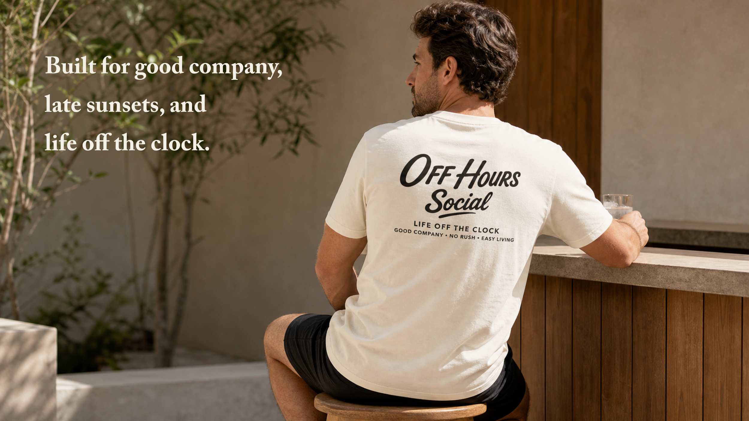 Man sitting on a wooden stool at an outdoor bar, wearing an off-white T-shirt with black text that reads "Off Hours Social" and sunglasses atop his head, holding a glass of water, with a background of tree branches and a beige wall.
