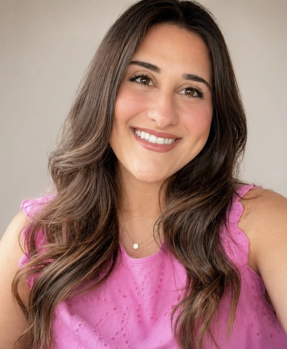 A smiling woman with long, wavy brown hair wearing a pink sleeveless top with eyelet details and a delicate necklace.