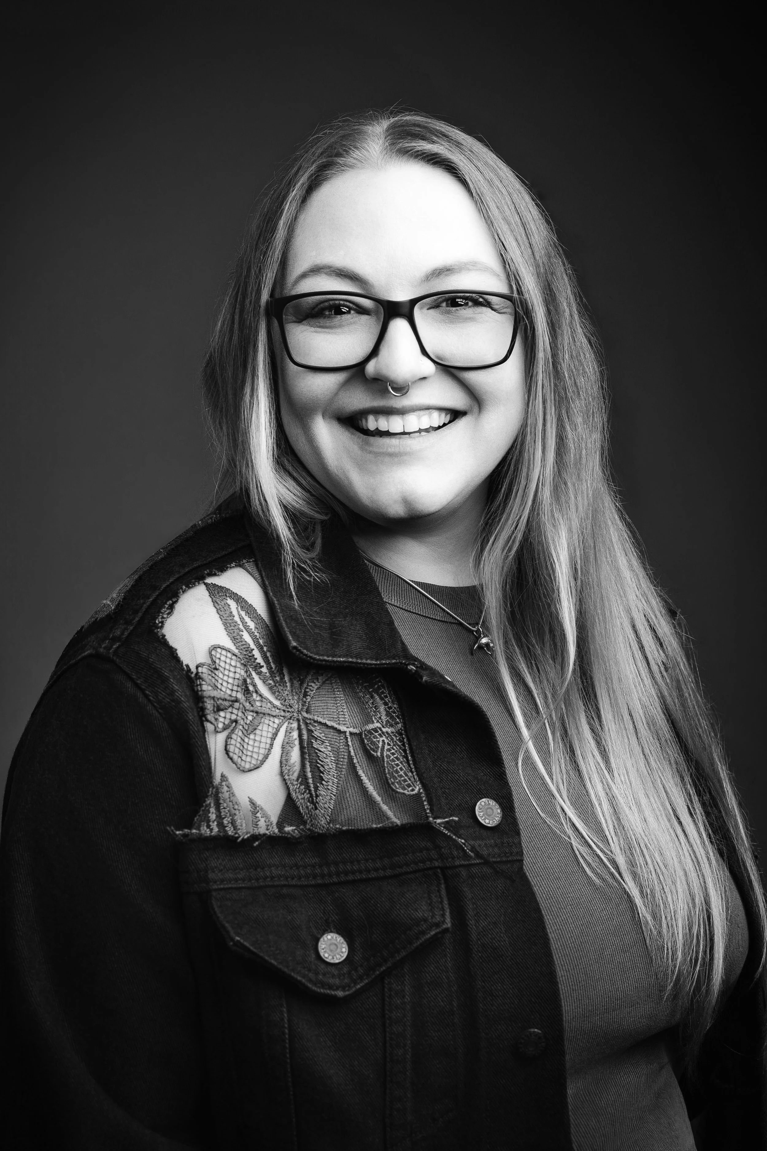 Black-and-white headshot of a smiling woman with glasses and long hair in a denim jacket with lace accents. Image by Adrianne Mathiowetz Photography