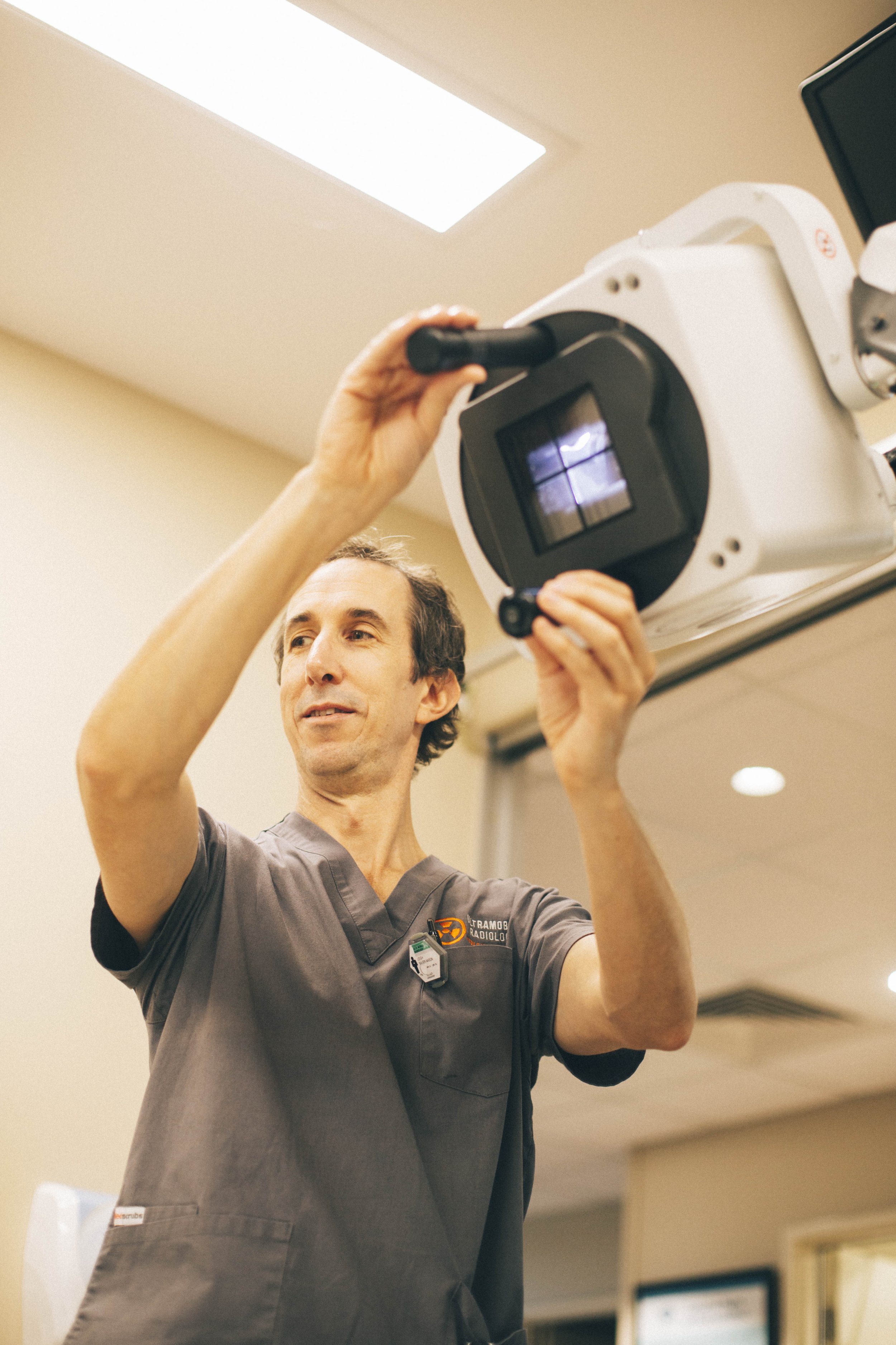 A male healthcare worker in gray scrubs, wearing an ID badge and a name badge, is adjusting a piece of medical imaging equipment in a medical setting.