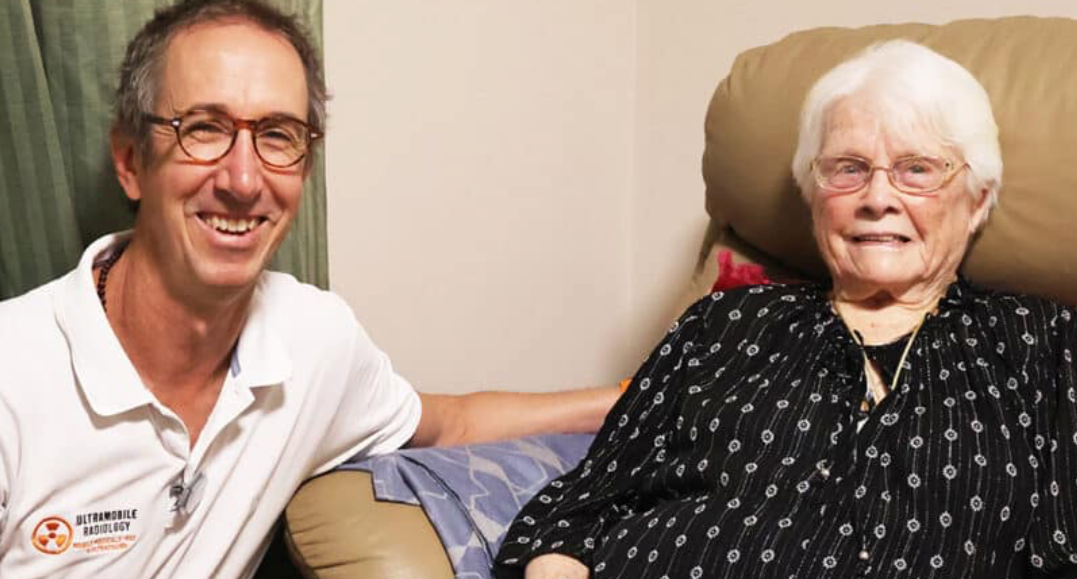 A man with glasses and a white polo shirt smiling and sitting next to an elderly woman with white hair and glasses, who is smiling and sitting in a recliner chair.