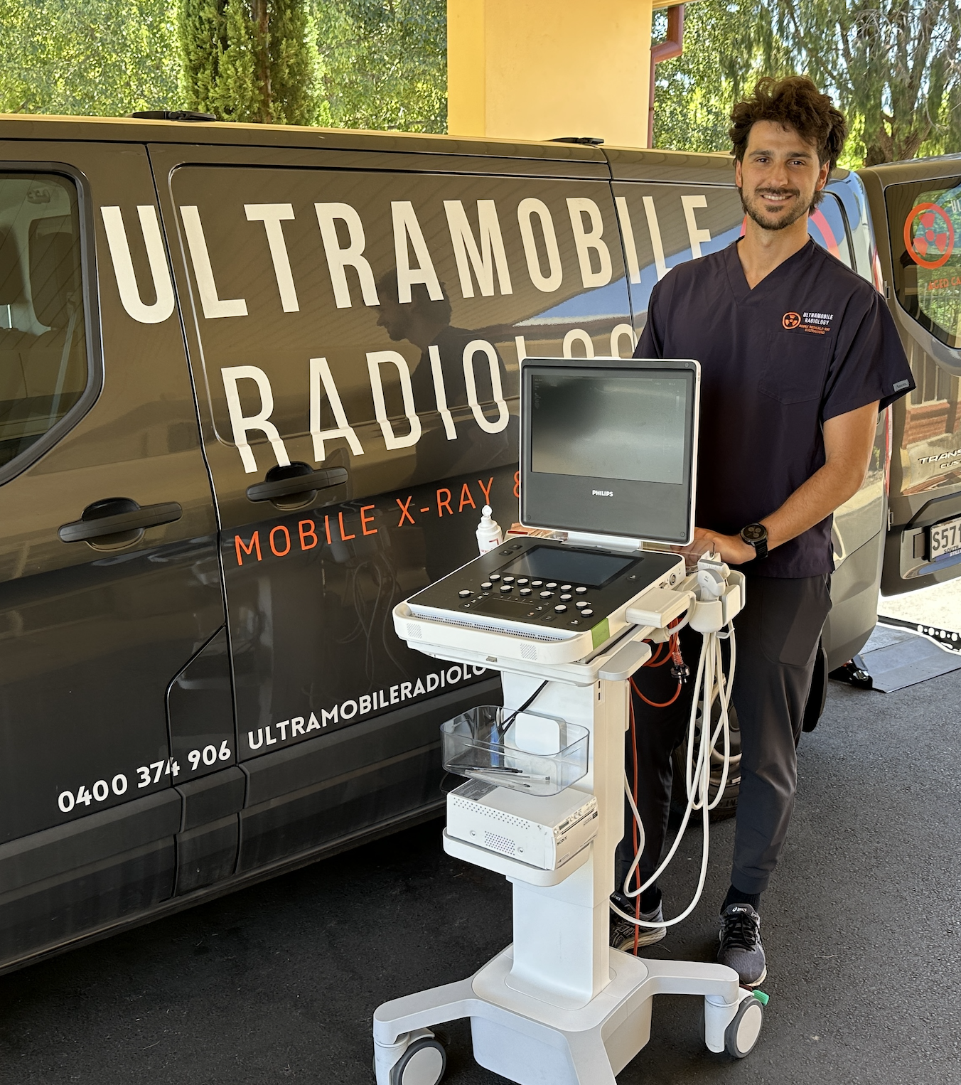 A male medical professional standing next to a mobile ultrasound device with a monitor, control panel, and attached probes, outside a van with 'UltratrMobile Radiology' written on it, during daytime with trees in the background.