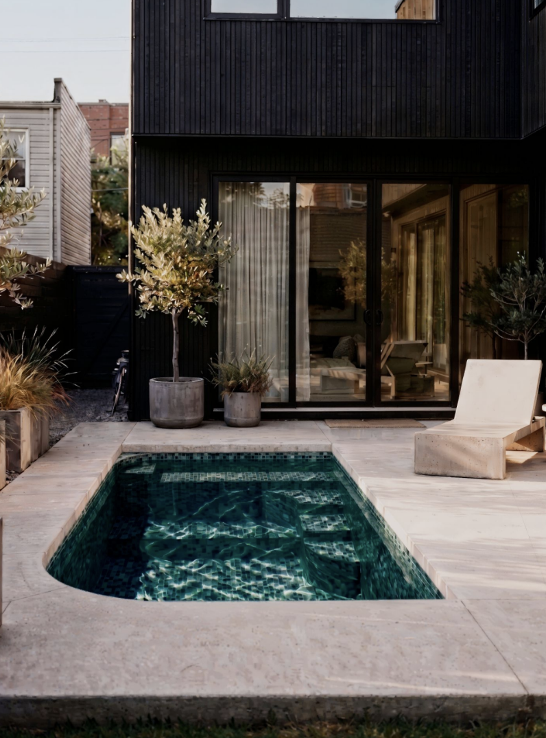 Modern backyard with a small rectangular pool, beige stone patio, potted plants, a white lounge chair, and a black house with large glass sliding doors.
