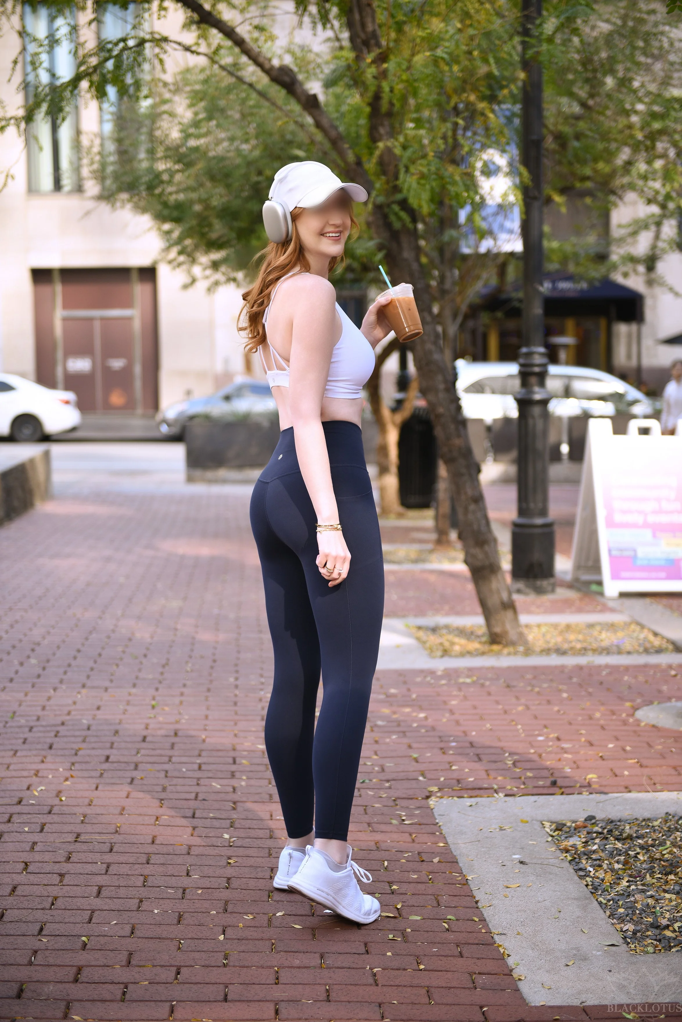 A woman with long, wavy brown hair is standing outside on a brick sidewalk, holding a plastic cup with iced coffee and a blue straw. She is wearing a white cap, large headphones, a white sleeveless top, black leggings, and white sneakers. There are trees and buildings in the background, and she is smiling at the camera.