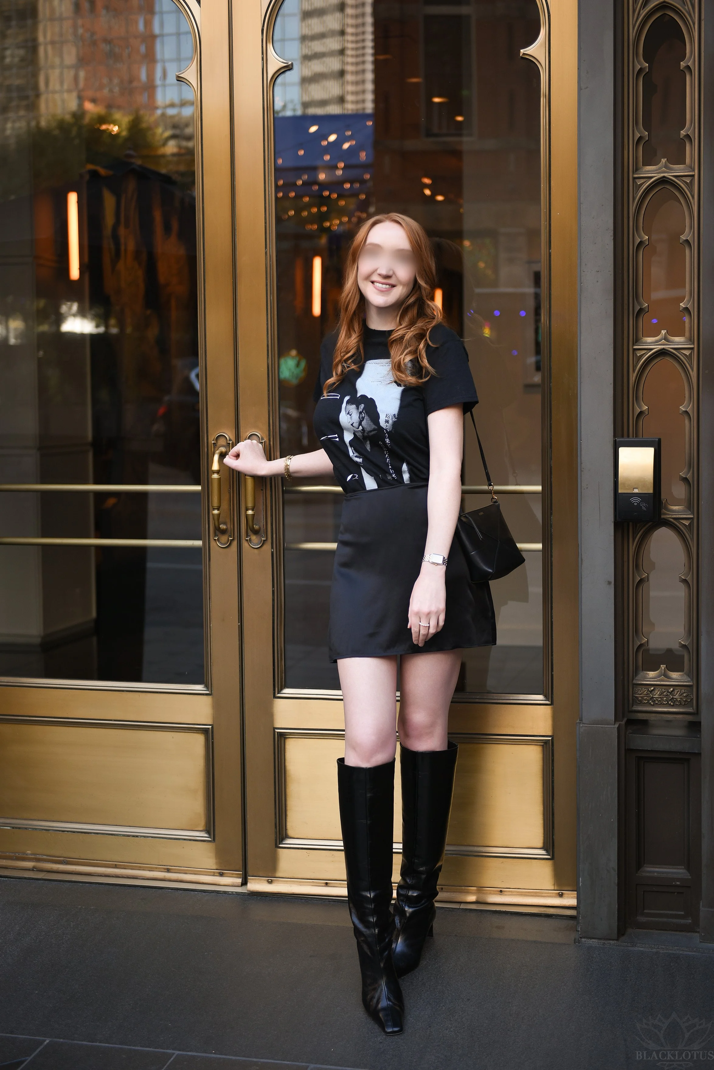 A woman with long wavy red hair standing in front of a large golden door with glass panels. She is smiling, wearing a black graphic T-shirt, a black skirt, and tall black boots. She has a black shoulder bag and some jewelry.