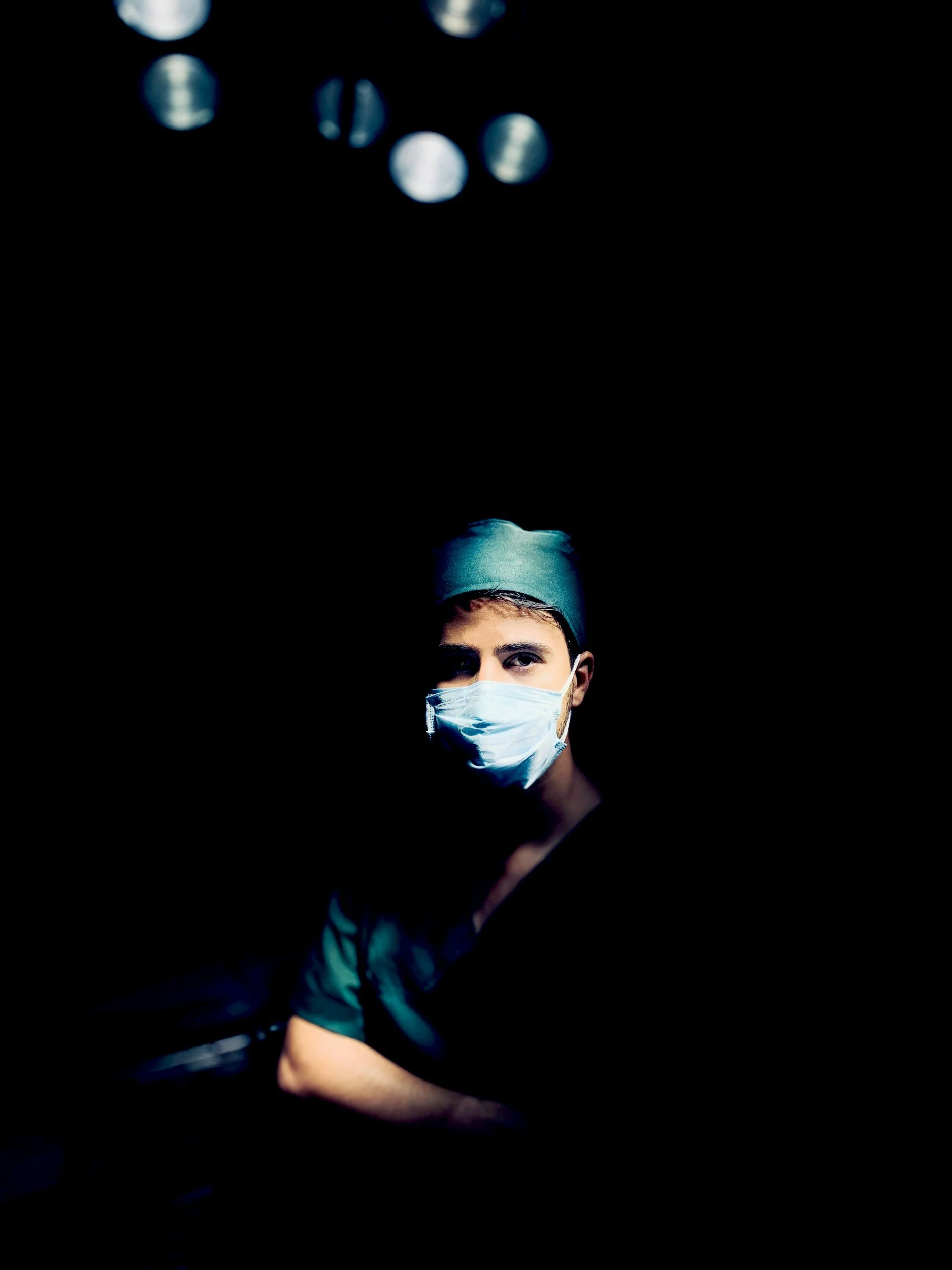 A person wearing a surgical mask and medical scrubs in a dark setting, illuminated by a light source from below, with their face partially visible.
