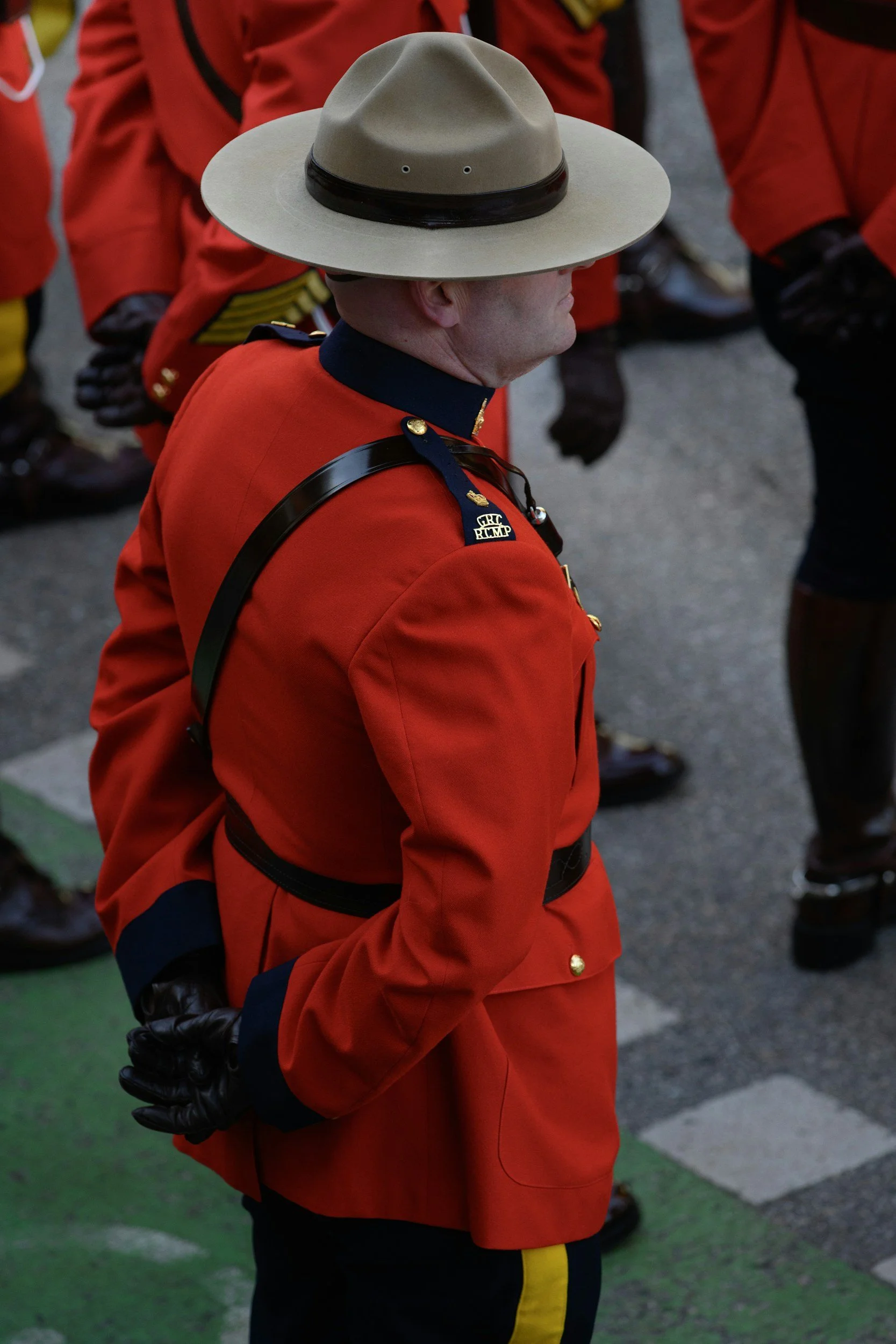 A man wearing a beige hat, red uniform, black gloves, and black pants with yellow stripe, standing with hands behind back during a parade or ceremony.