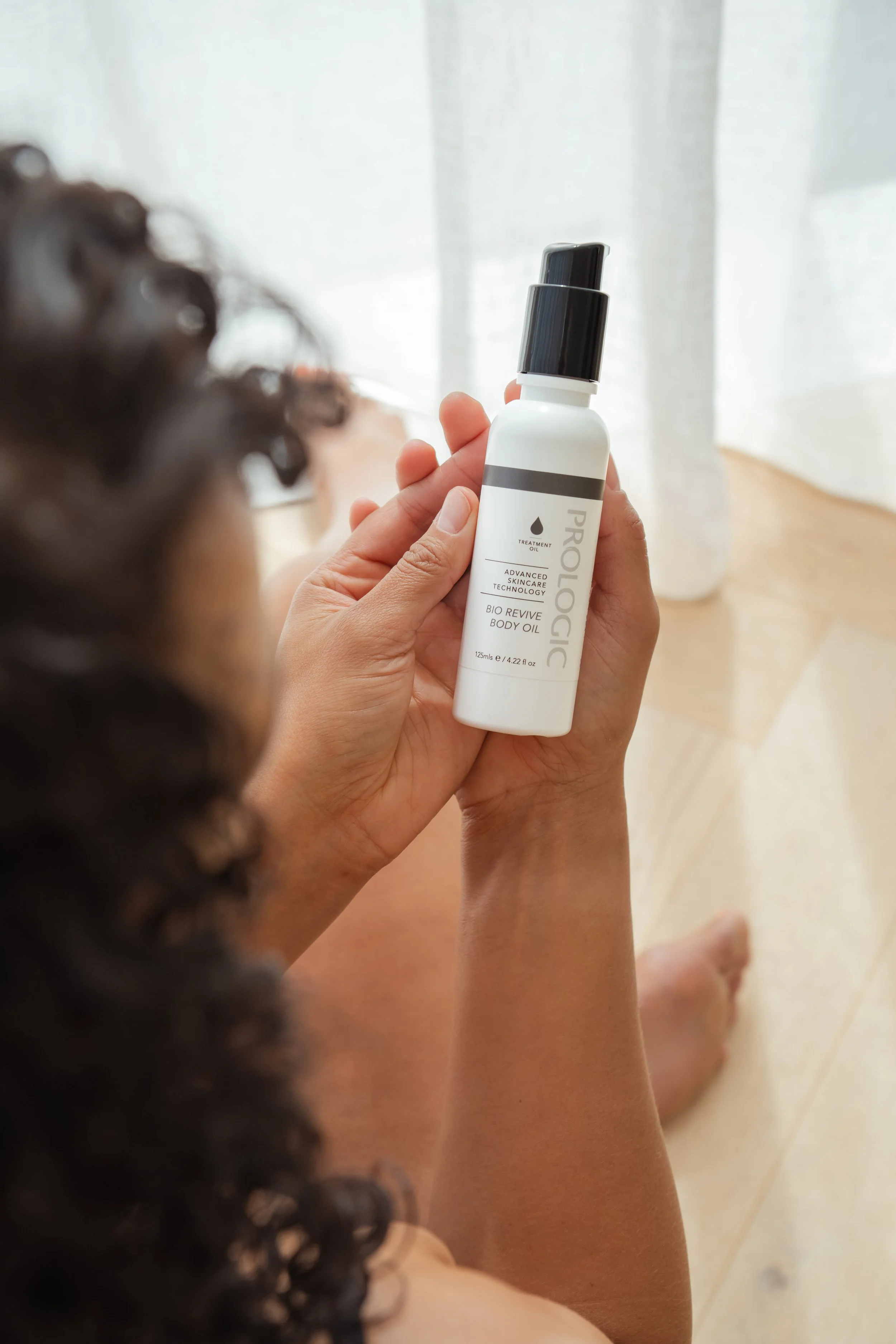 A person with curly hair holding a white bottle labeled 'Prológica Bio Revive Body Oil' with a black pump dispenser near sheer curtains.