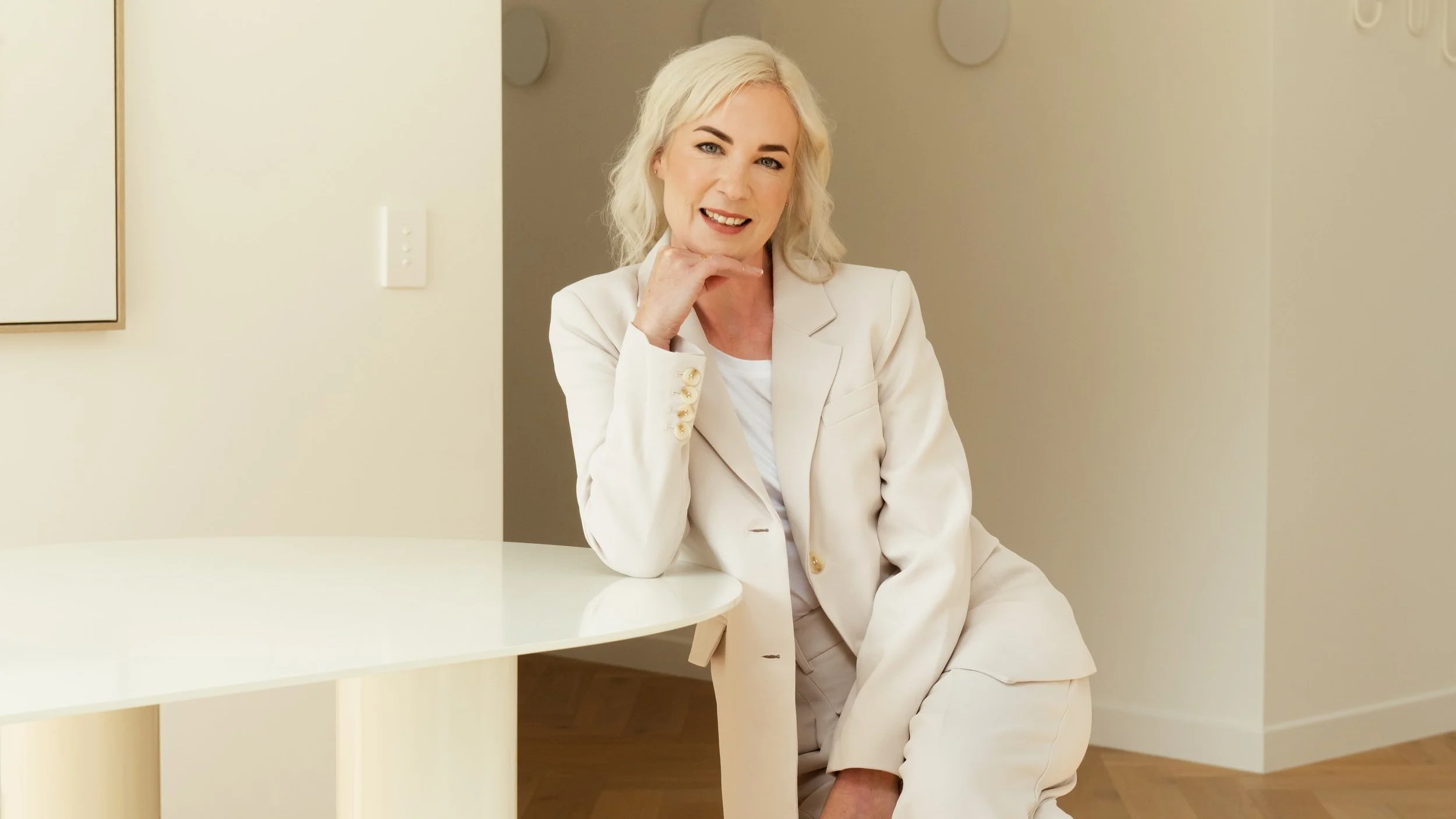 A smiling woman with blonde hair in a white blazer and pants, sitting and leaning on her hand, in a neutral-colored room.