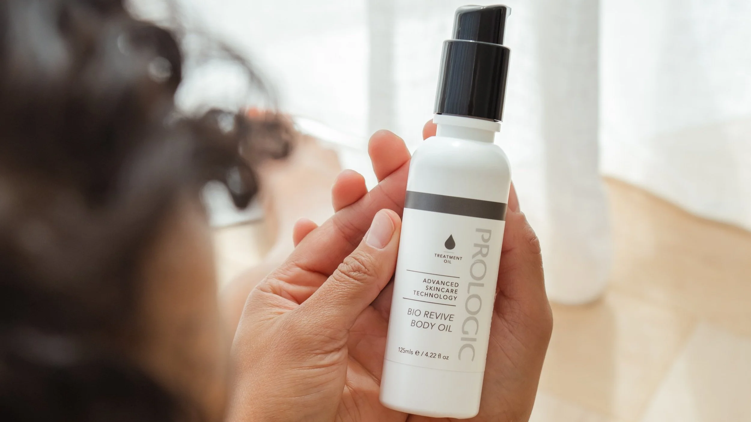 Close-up of a person holding a white bottle of skincare or body oil with a black cap, with a blurred background and the person's face out of focus.