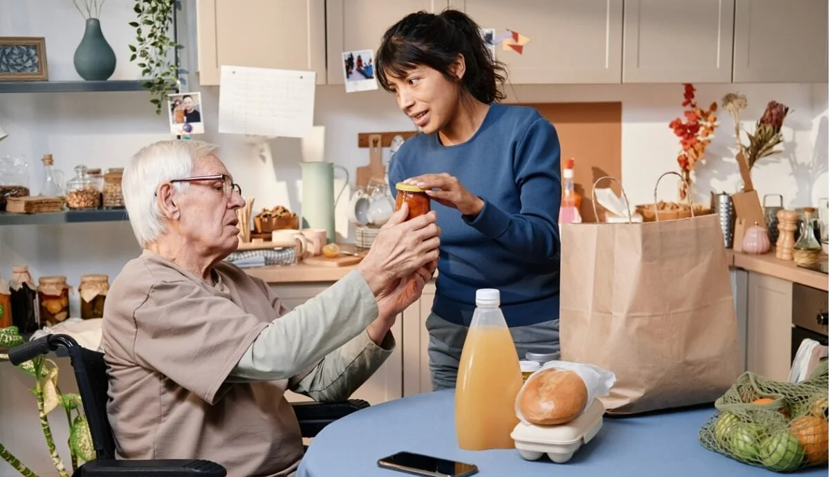 A young woman is giving a jar of jam or honey to an elderly man in a wheelchair in a kitchen.