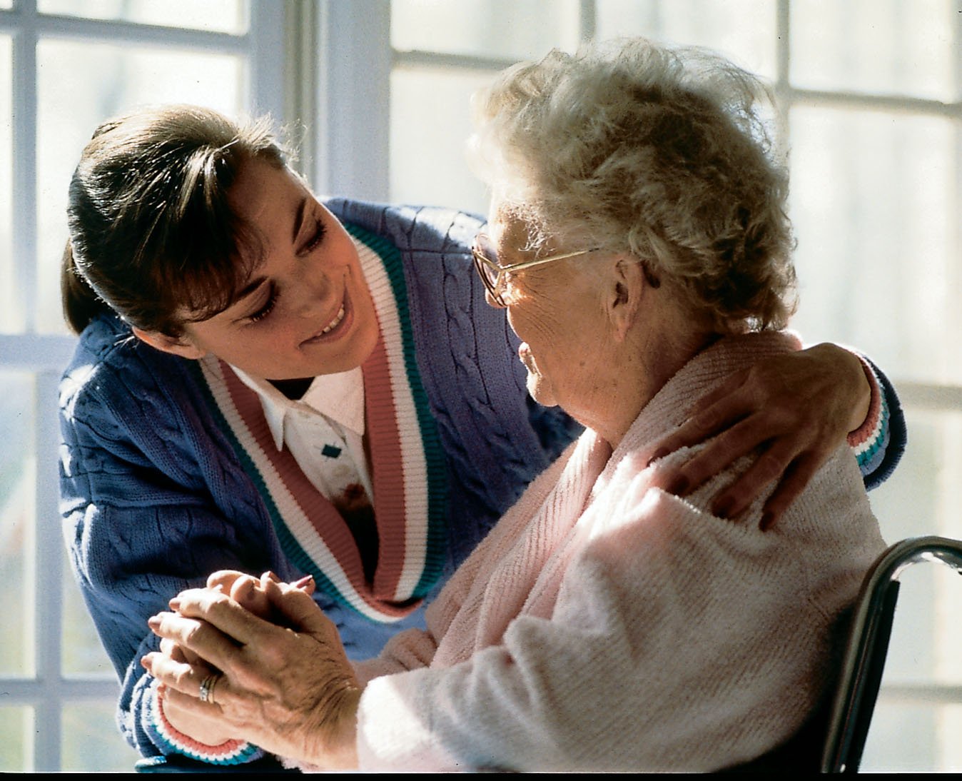 A young woman is smiling and holding hands with an elderly woman who is sitting in a wheelchair. The young woman has dark hair and is wearing a colorful sweater, while the elderly woman has gray hair, glasses, and is wearing a light peach sweater. They are near a window with sunlight streaming in.
