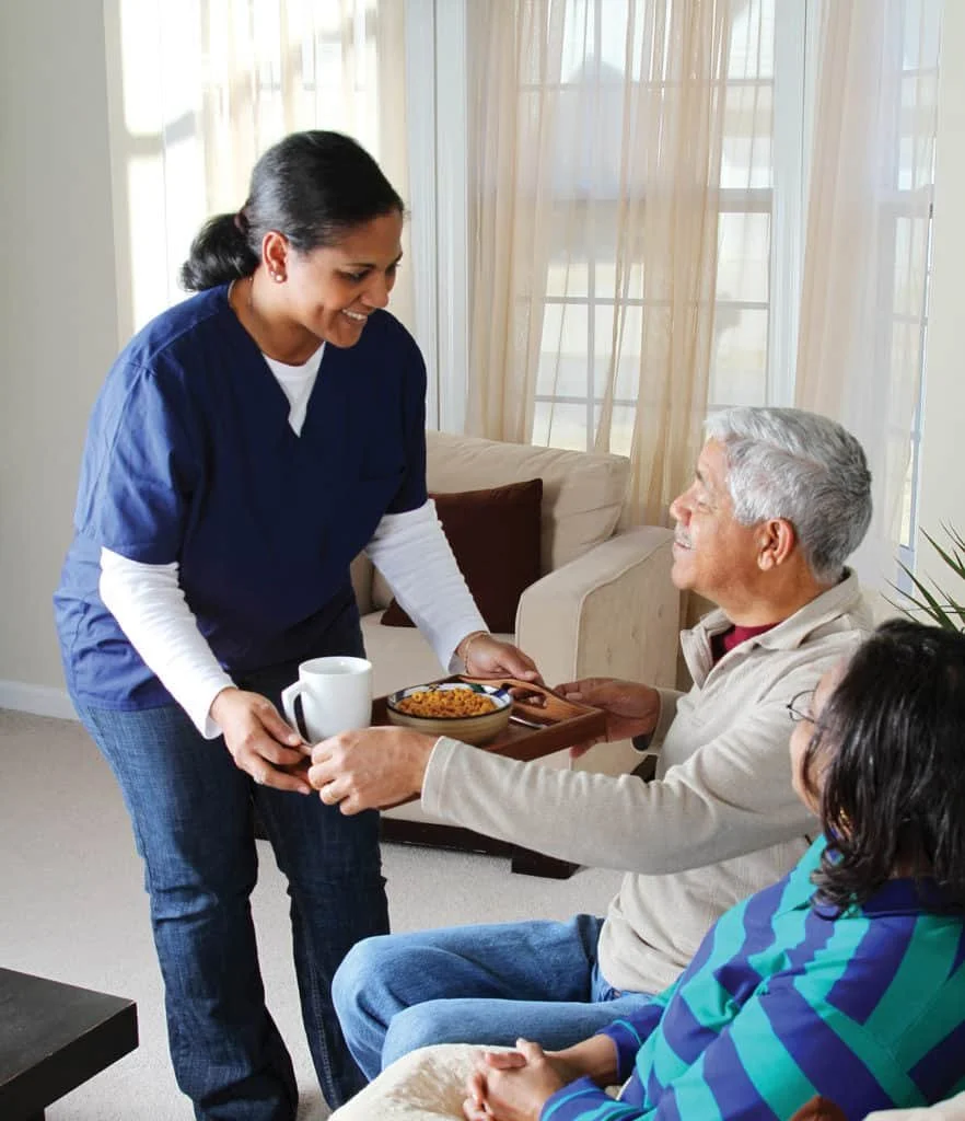 A caregiver hands a tray with food and a cup to an elderly man sitting on a sofa, with a woman sitting beside him, in a bright living room.