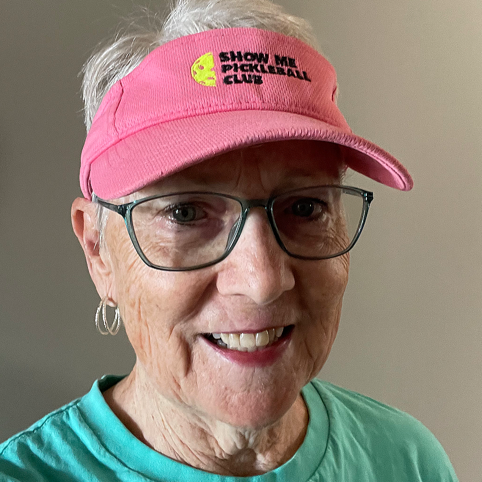 An older woman wearing glasses and a pink visor hat that says 'Show Me Pickleball Club' with a yellow pickleball logo, smiling at the camera.