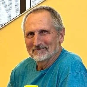 An older man with gray hair and beard, wearing a blue shirt, sitting indoors near a yellow wall.