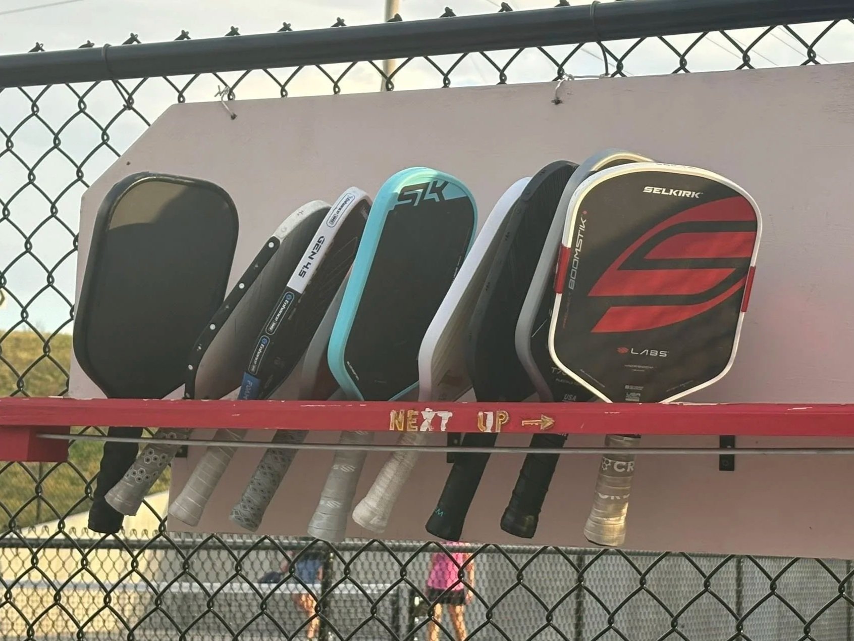 Pickleball paddles placed on a rack at a fence-lined pickleball court